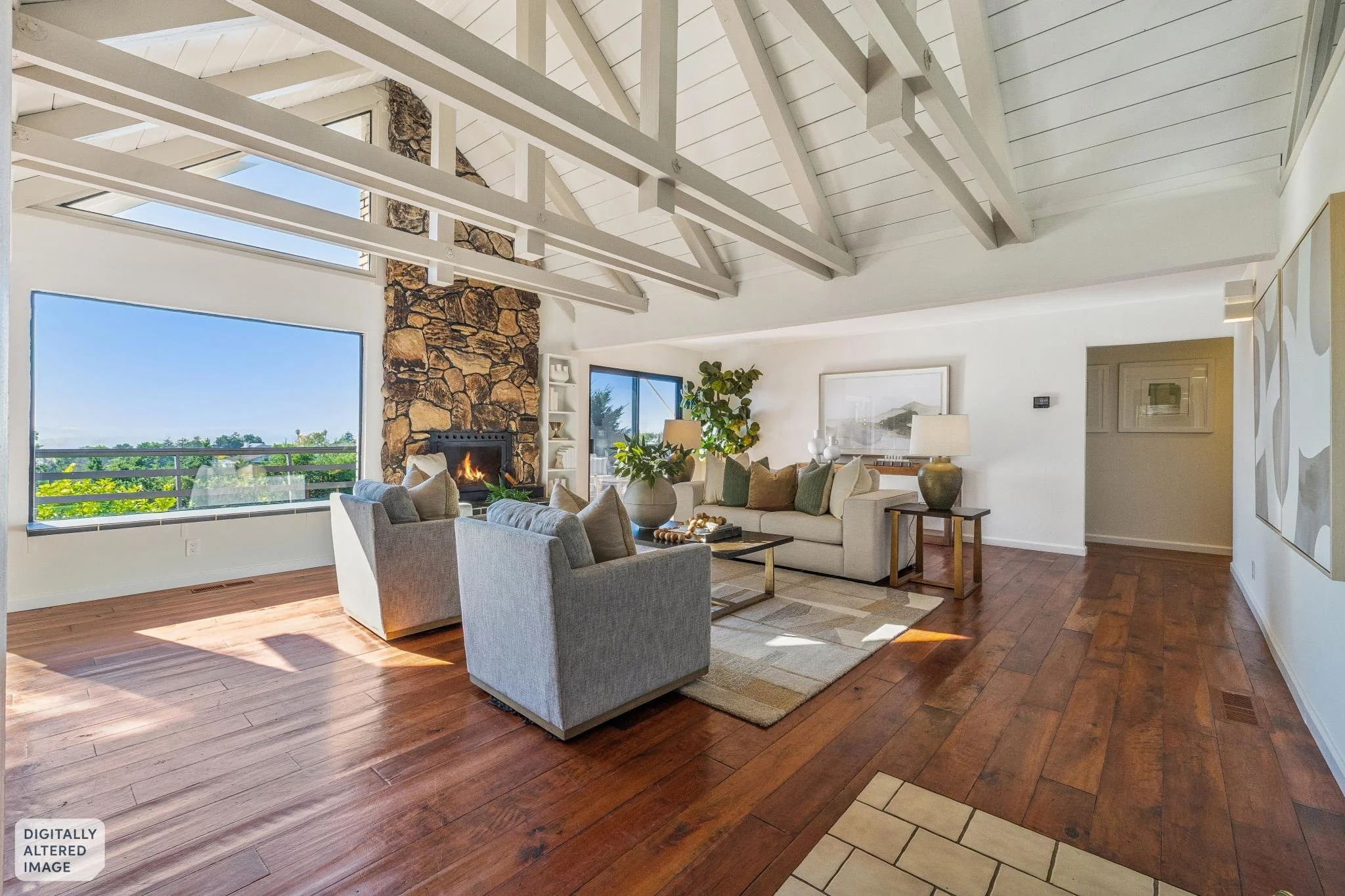 Interior view of a spacious living room with white walls, wooden flooring, large windows, a stone fireplace, and modern furniture.