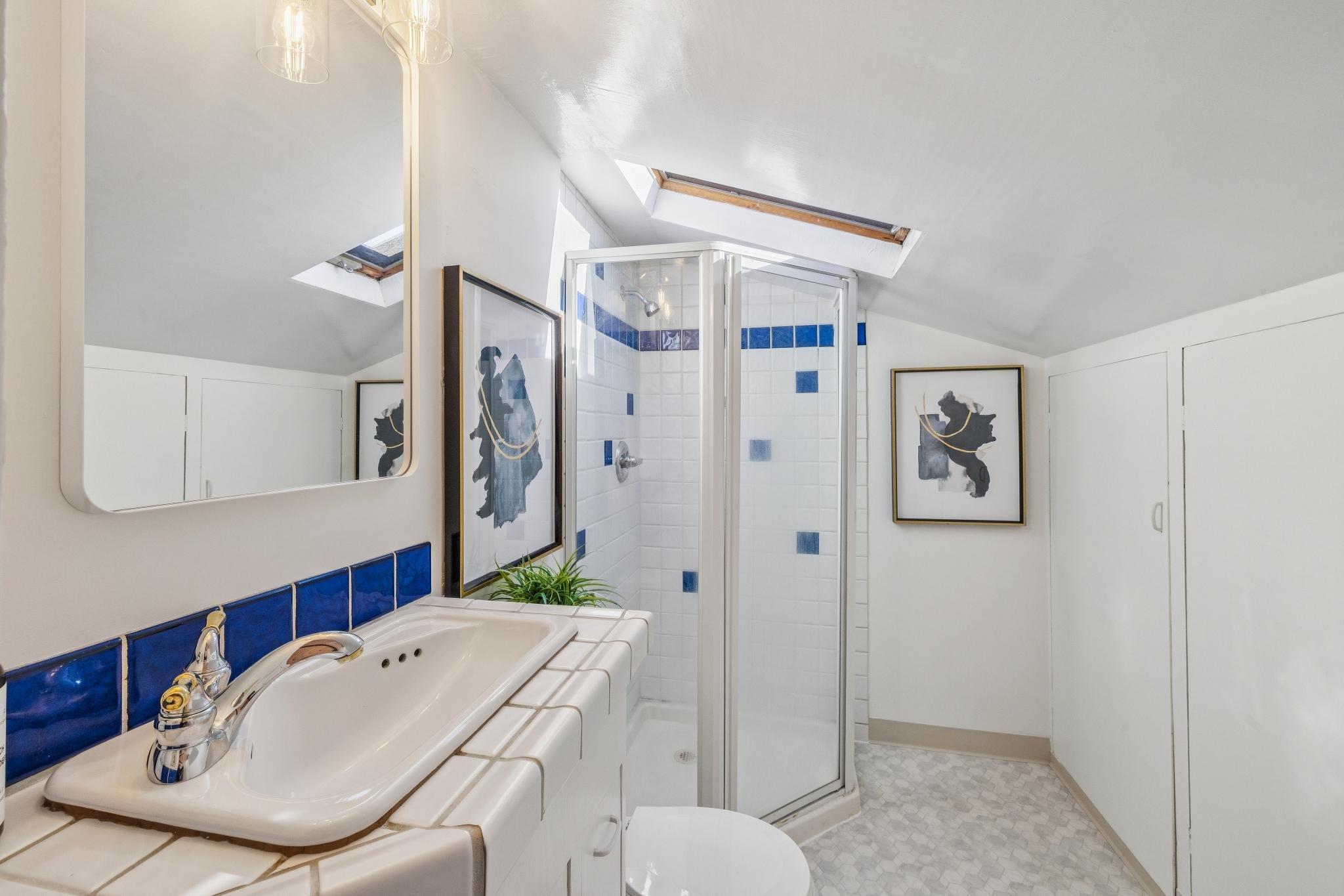 A bathroom with a white tile sink, blue tile accents, a mirror, artwork of animals, a glass shower, and skylight windows.