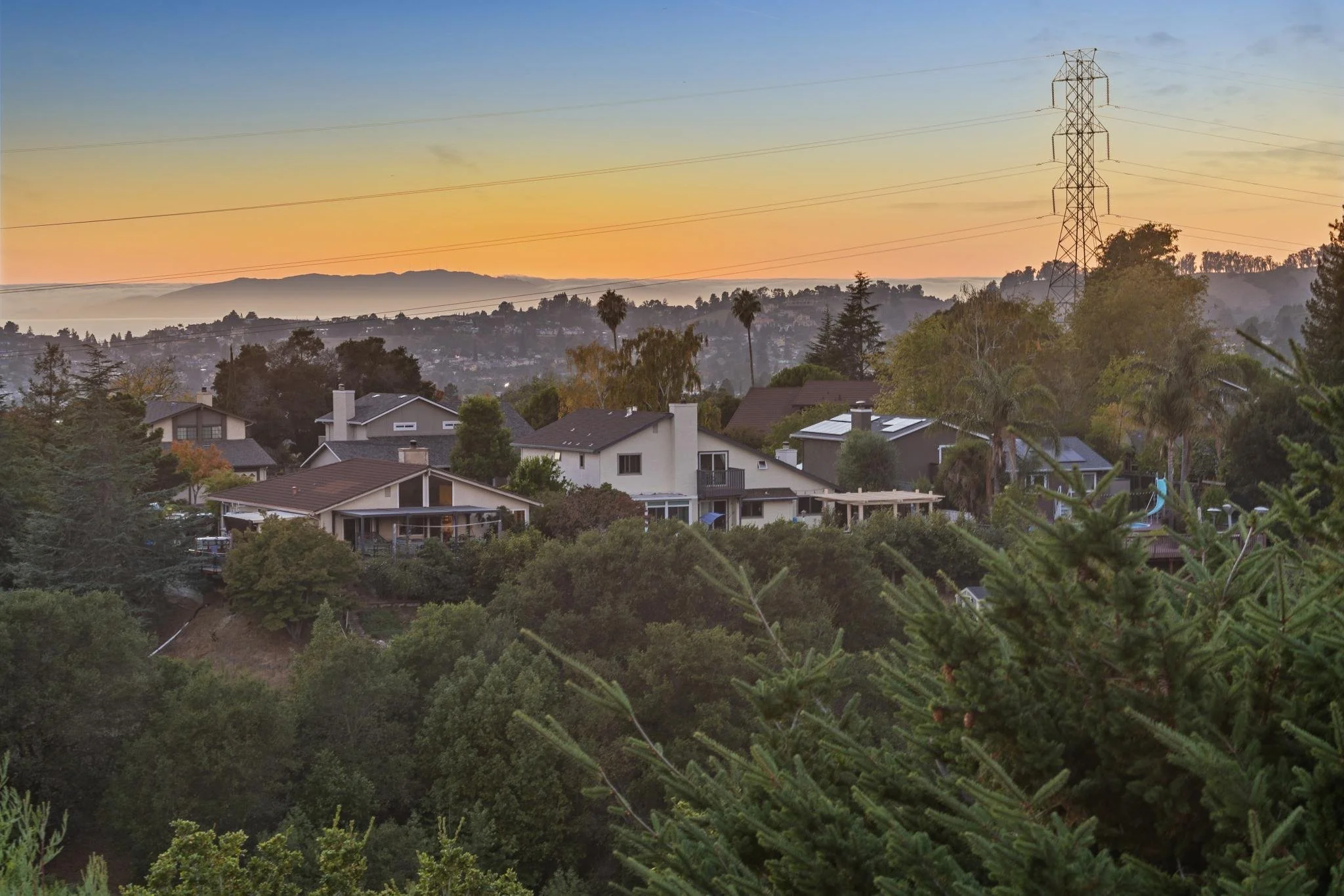 Residential neighborhood with houses surrounded by trees, hills in the background, and a large electrical transmission tower, all under a sunset sky.