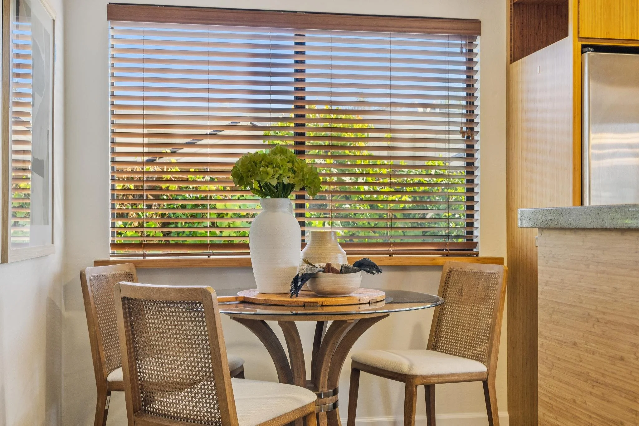 A dining table with four chairs and a large vase with green hydrangea flowers, positioned in front of a window with wooden blinds, in a bright room.