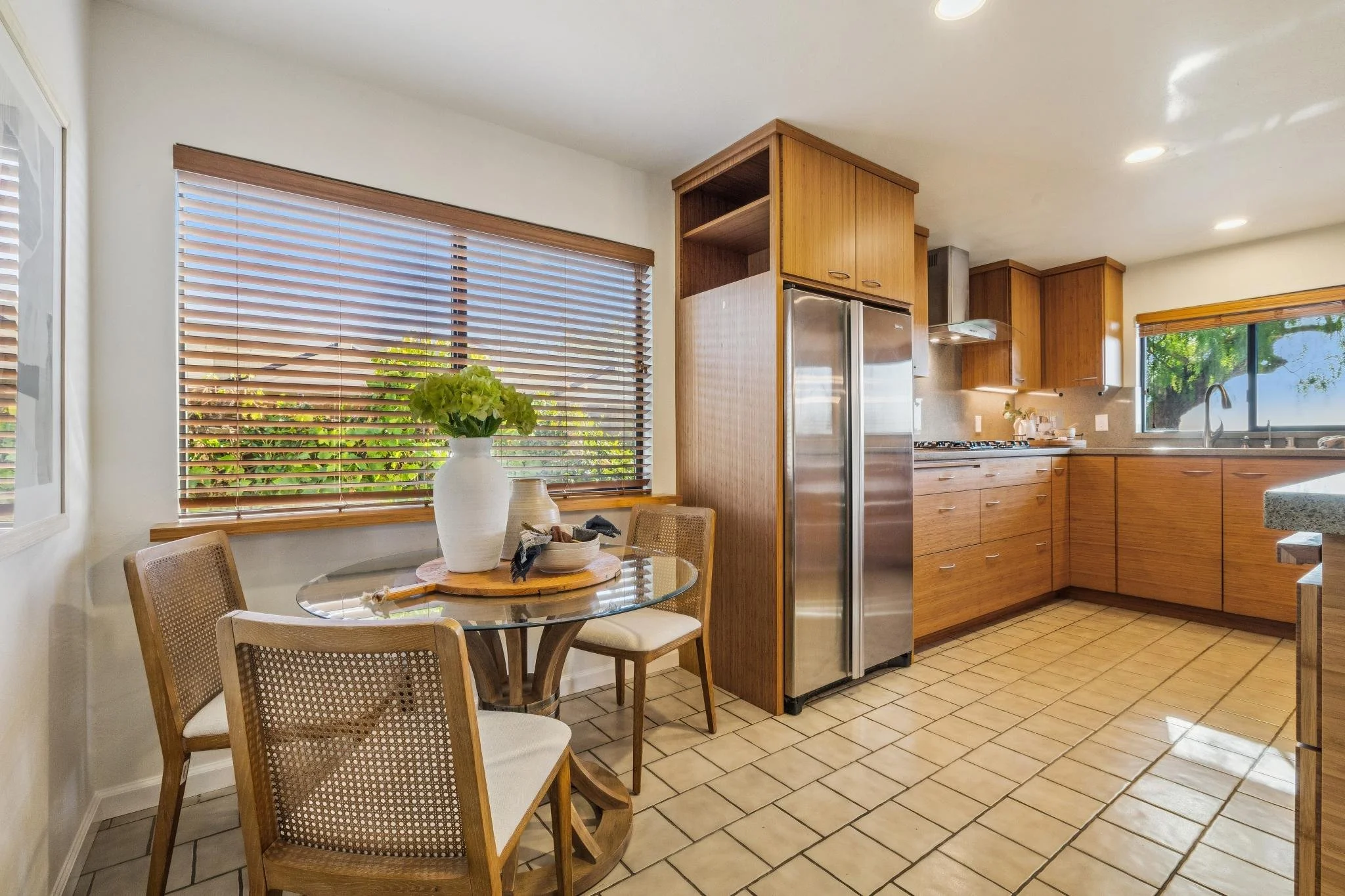A kitchen featuring wooden cabinets, a stainless steel refrigerator, a tiled floor, a window with wooden blinds, and a round dining table with four chairs and a white vase with green flowers.