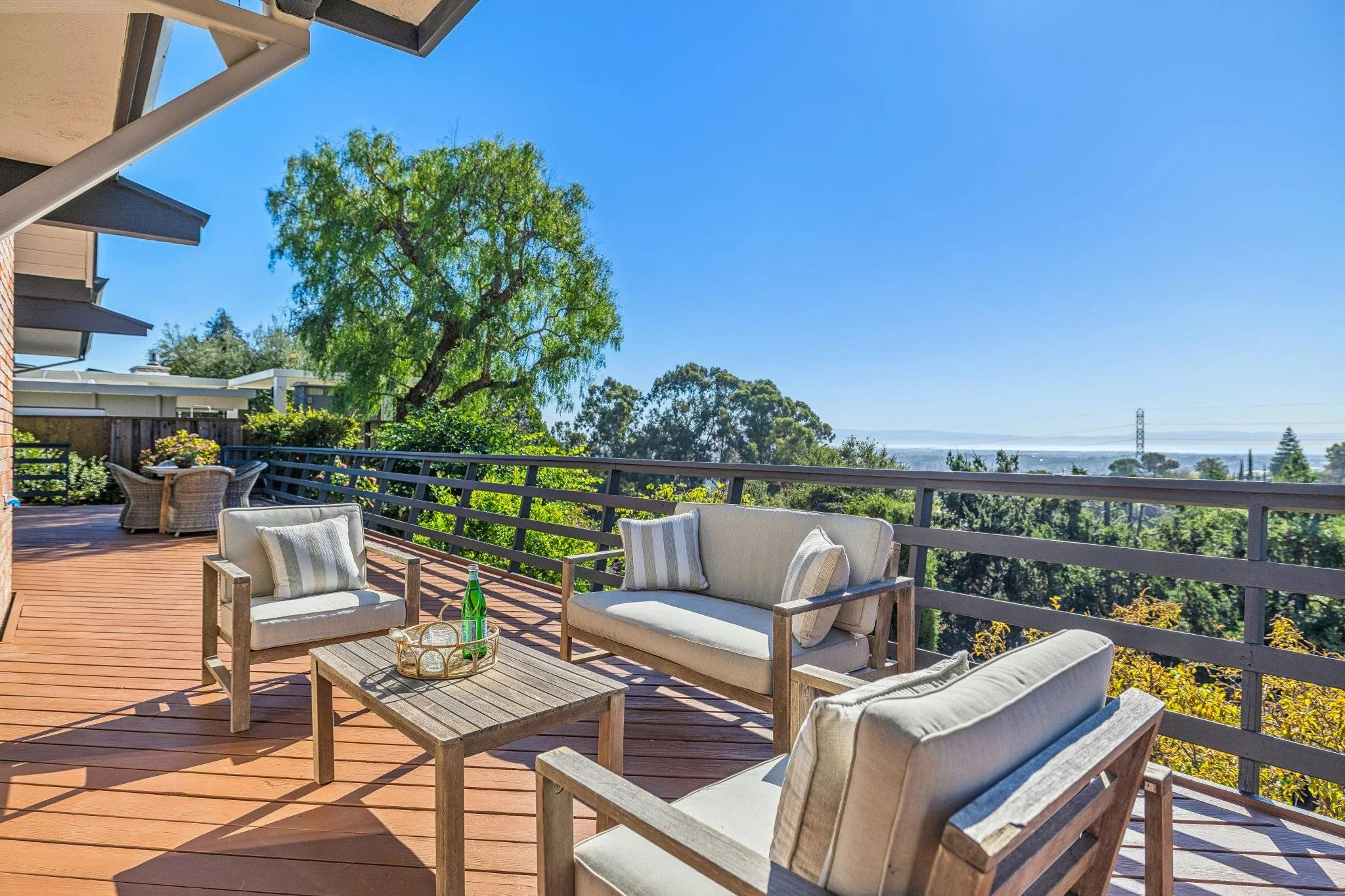 Outdoor balcony with wooden furniture, including a sofa, armchairs, and a coffee table, overlooking trees and a cityscape in the distance on a clear sunny day.