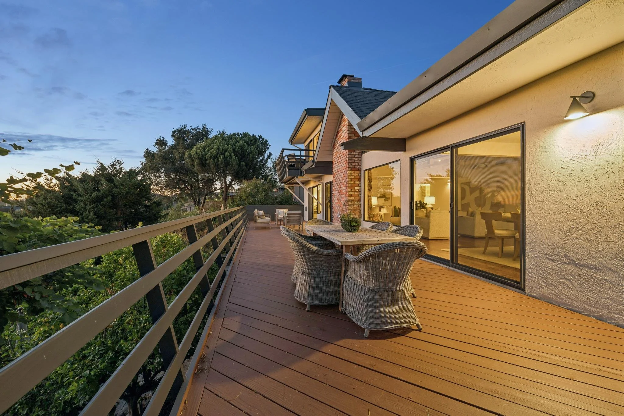 Residential balcony with wooden deck, outdoor wicker furniture, string lights, large sliding glass door, trees, and neighboring houses at sunset.