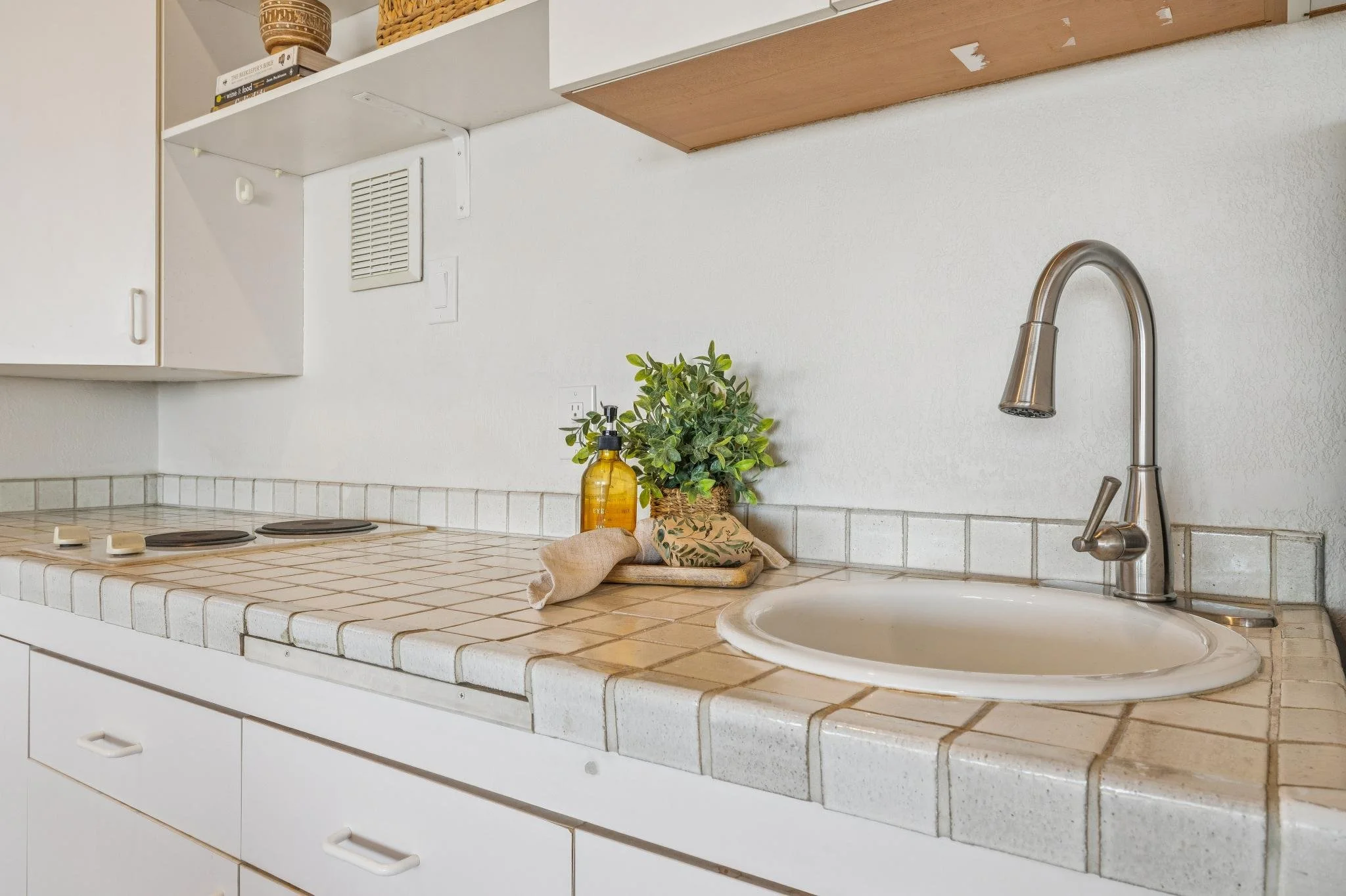 A kitchen countertop with a white tiled surface, a stainless steel sink with a curved faucet, a small potted green plant, a yellow soap dispenser, and a cloth along with other kitchen items. Above the countertop, there are white cabinets and a few de