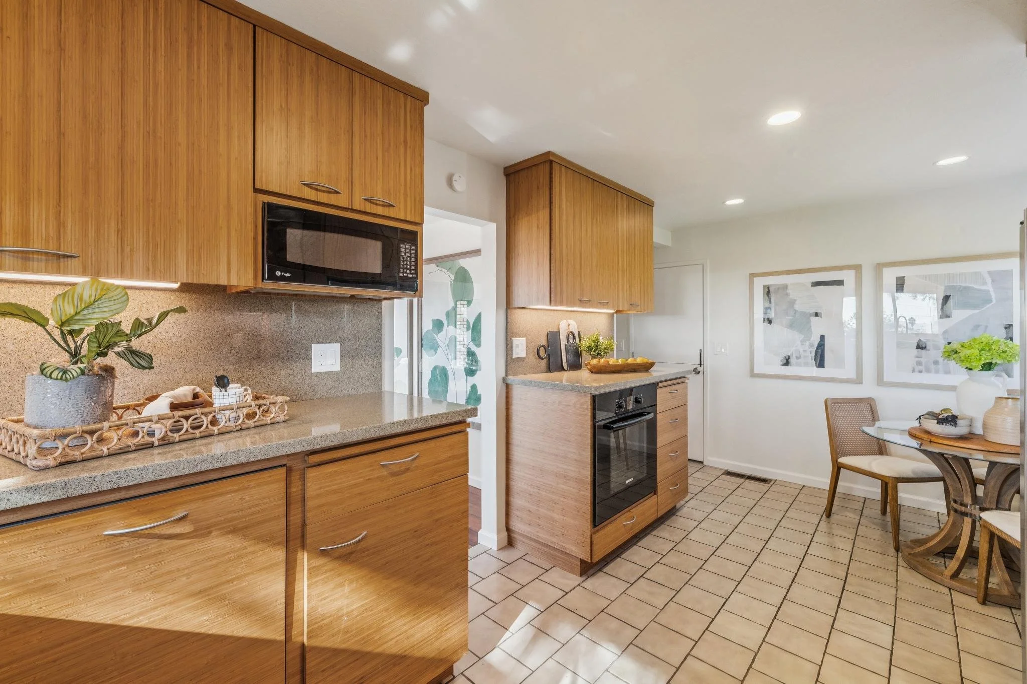A kitchen with wooden cabinets, a countertop with a potted plant, a microwave, a stove, and a dining area with a round table and chairs, large windows, and framed artwork on the wall.