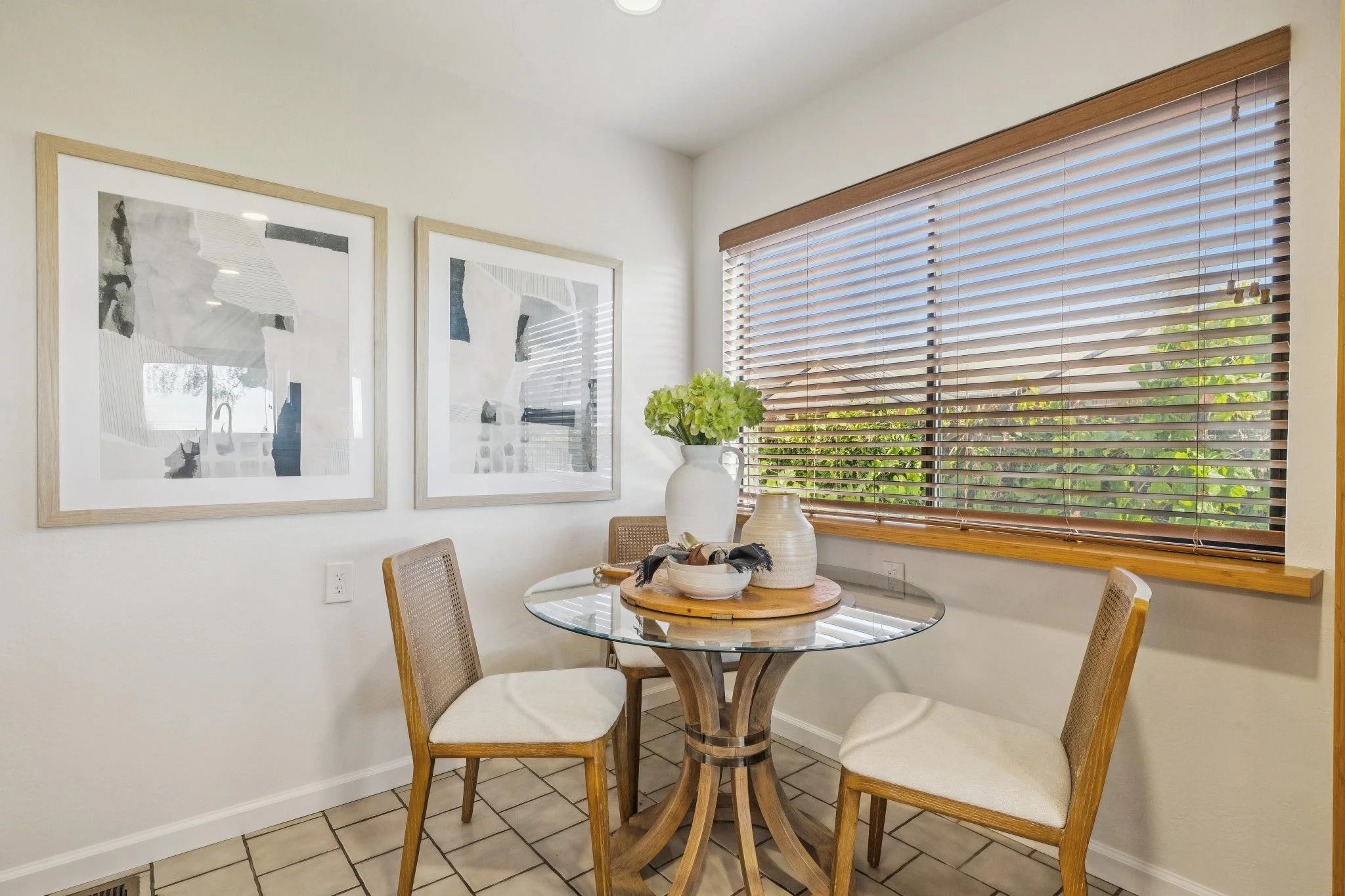 A kitchen corner with a round glass table, four wooden chairs with cushioned seats, a large white vase with green hydrangeas, and two black and white abstract art pieces on the wall. There are wooden blinds on a large window allowing sunlight to ente