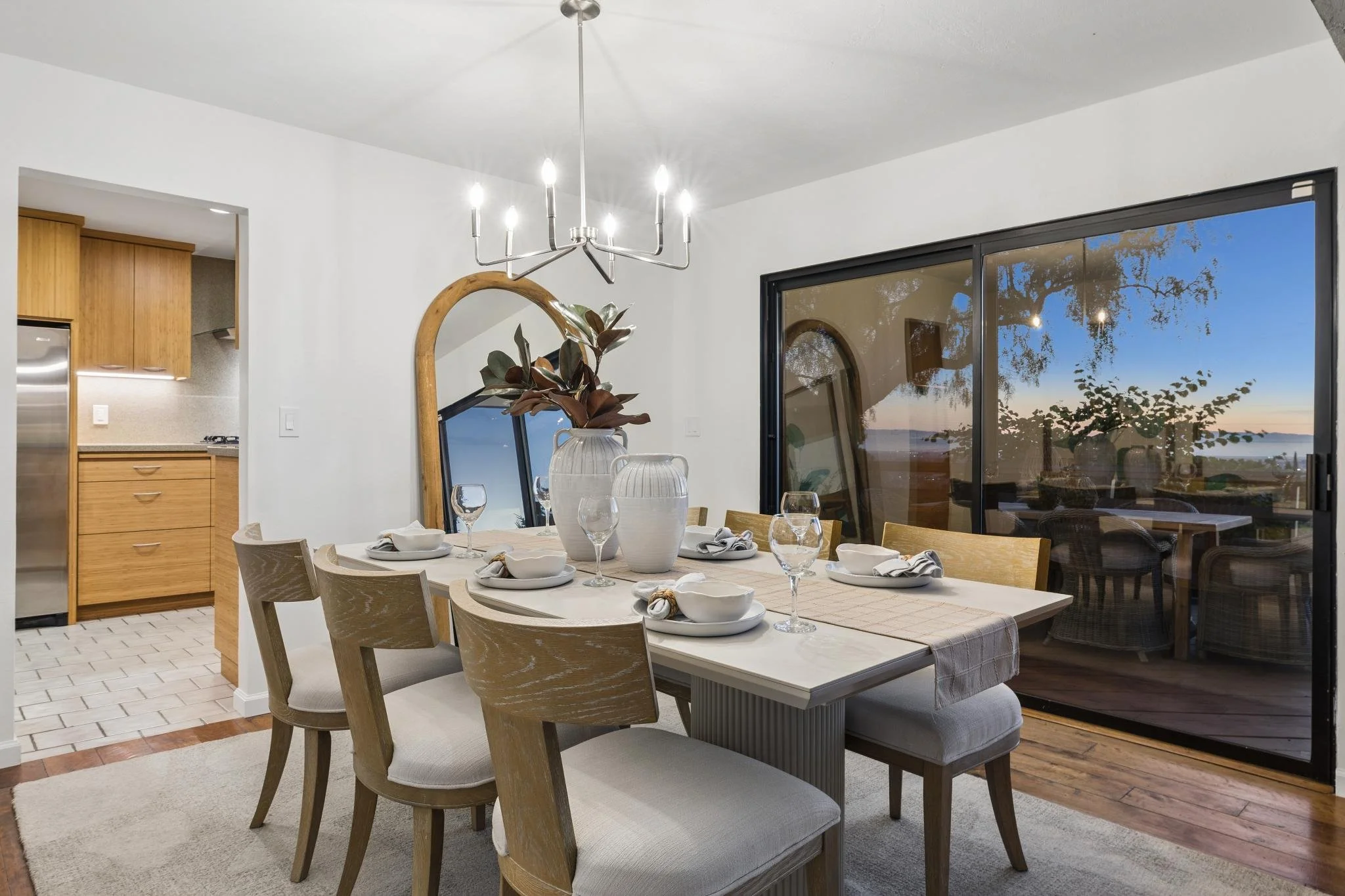 A dining room with a table set for six, featuring white bowls, glasses, and napkins, with a large window revealing a scenic ocean view at sunset.