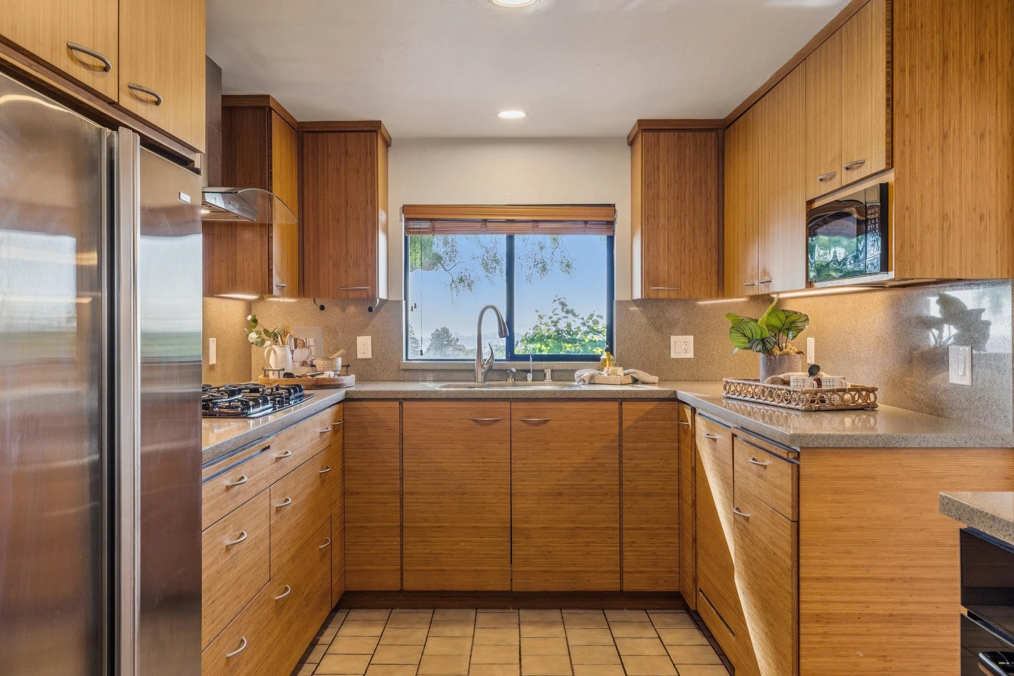 A kitchen with wooden cabinets, a window above the sink, a stainless steel refrigerator, a gas stove, and various decorative plants and items on the countertop.