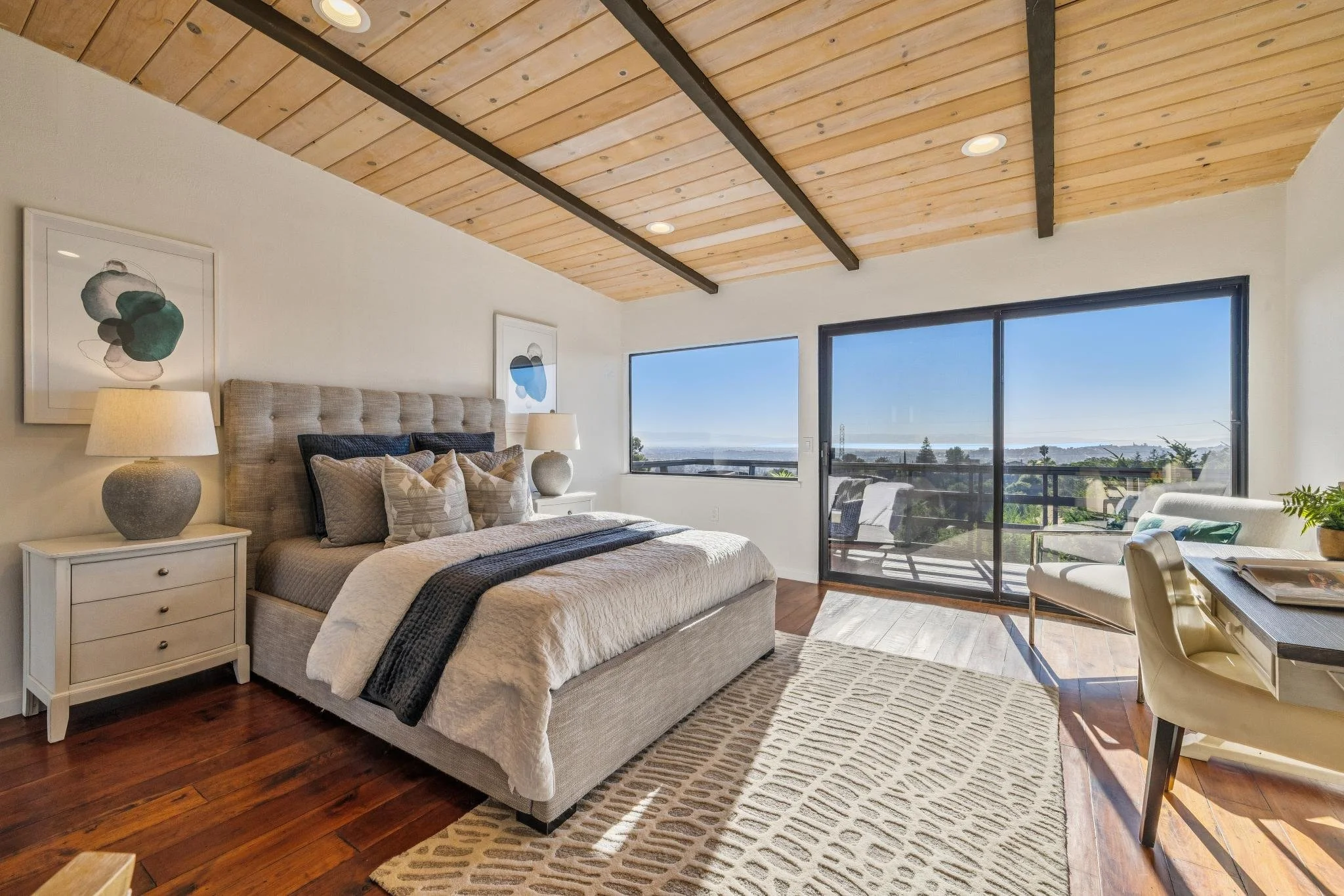 Bright bedroom with a wooden ceiling, large windows, a bed with beige and dark pillows, two nightstands with lamps, artwork on the walls, a chair, a desk, and an outdoor balcony with a view of trees and the ocean.
