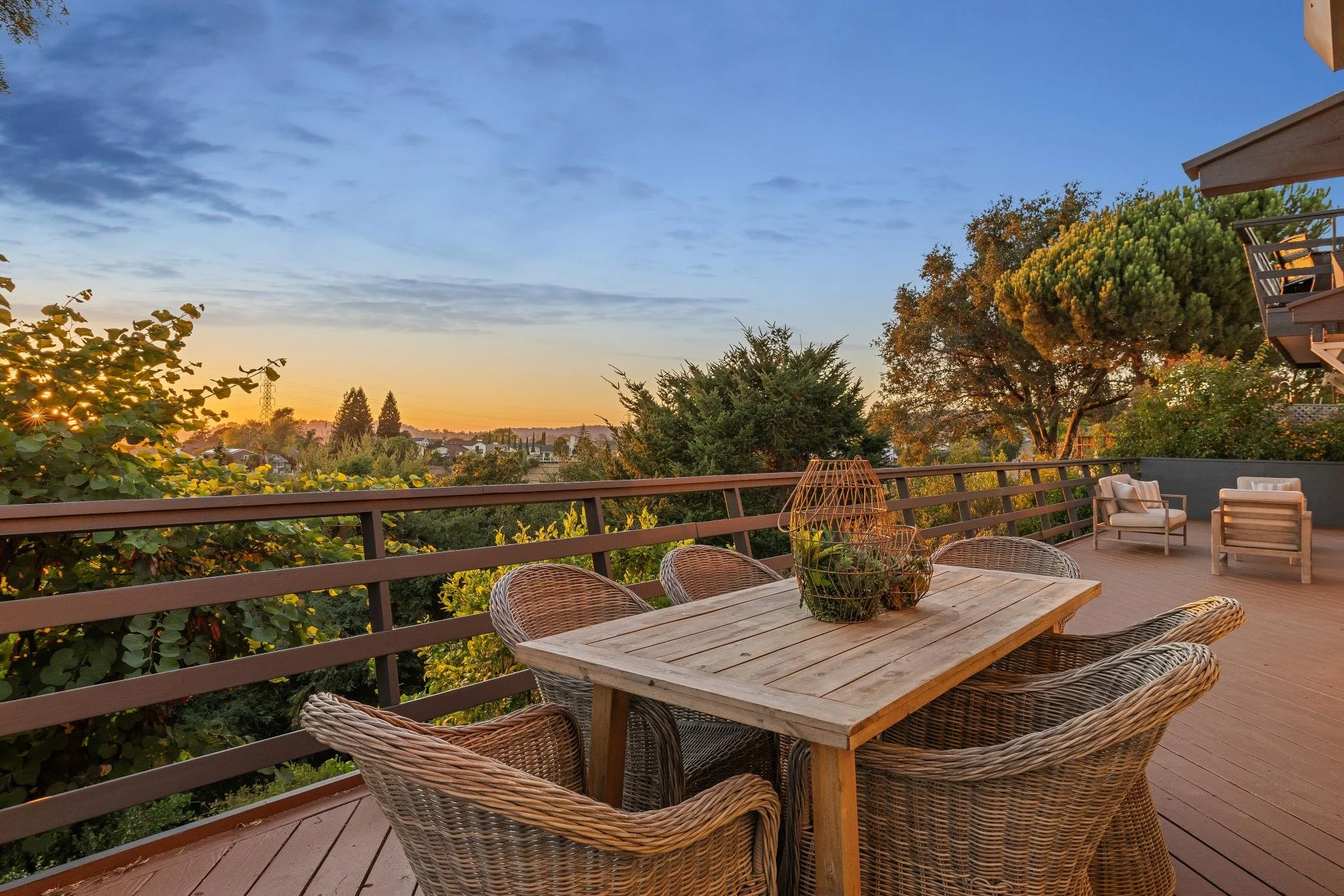 An outdoor balcony patio with a wooden table and wicker chairs, overlooking trees and a sunset sky with clouds and a neighborhood in the distance.