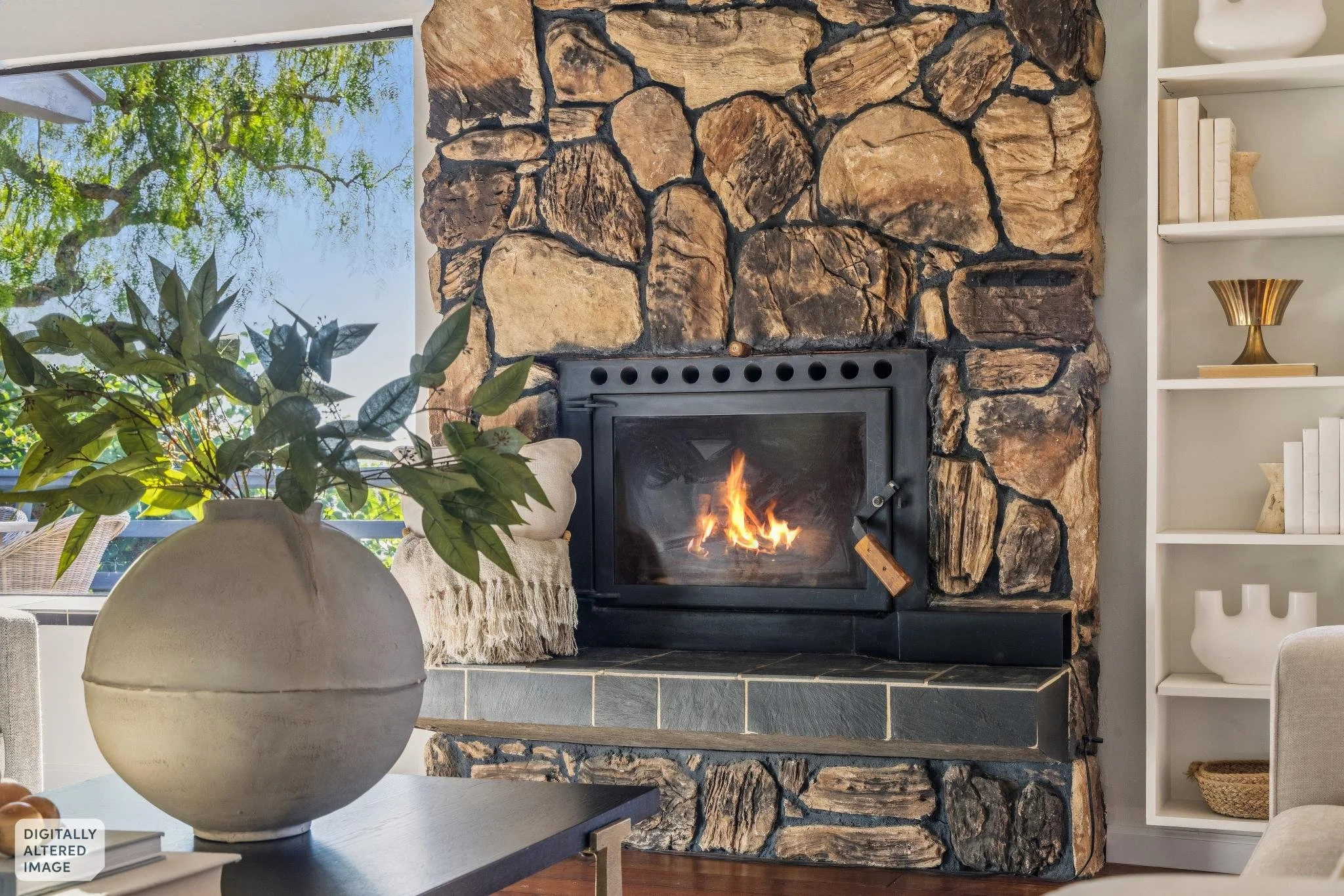 Living room with a stone fireplace and a fire. There is a large window with trees outside, a white bookshelf with books and decorative objects, a gold vase on a shelf, a beige armchair with a throw blanket, a large potted plant near the window, and a dark table with books and a large ceramic vase in the foreground.