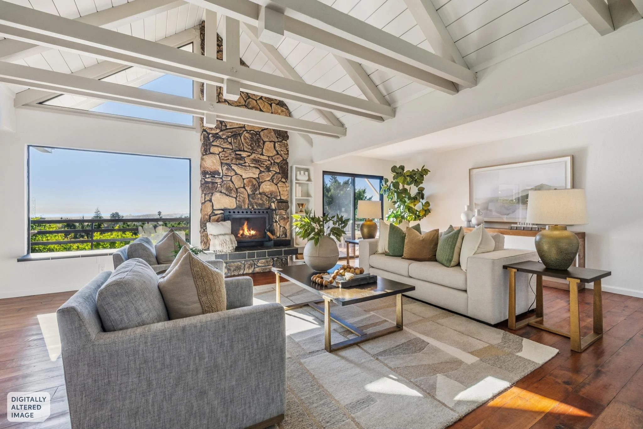 Living room with white walls, wooden floor, large windows, a fireplace with a stone surround, beige sofas, a gray armchair, wooden side tables with lamps, green plants, and decorative items.