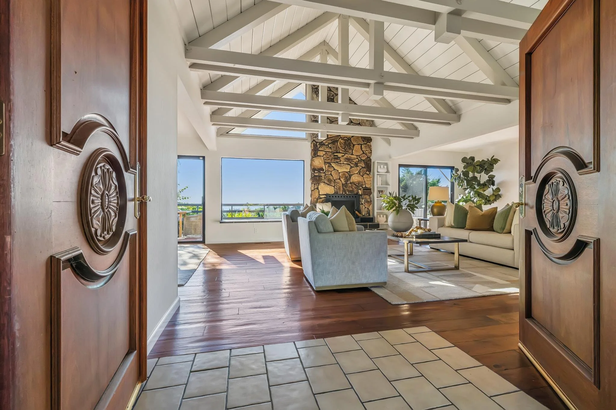 An open front door leading into a spacious living room with vaulted ceilings, exposed beams, large windows, a stone fireplace, and modern furniture