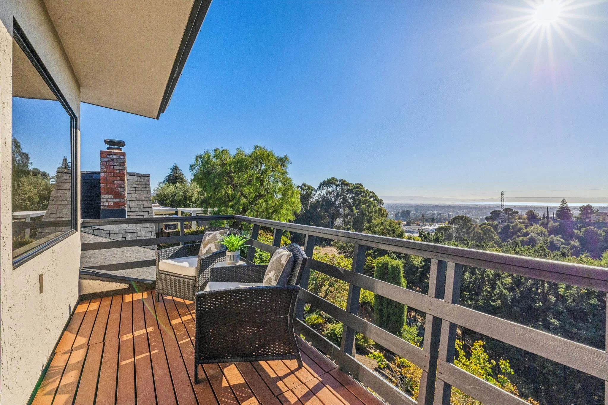 Balcony with outdoor furniture, including a table and chairs, overlooking a scenic view of trees, hills, and a cityscape in the distance under a clear blue sky with the sun shining.
