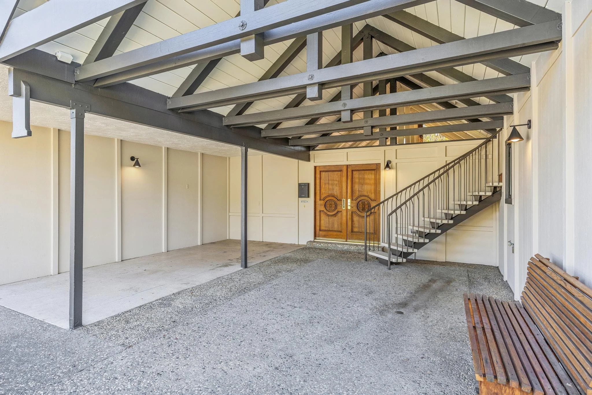 Covered parking area with a concrete floor, beige walls, wooden double doors, staircase with metal railing, and wooden benches.