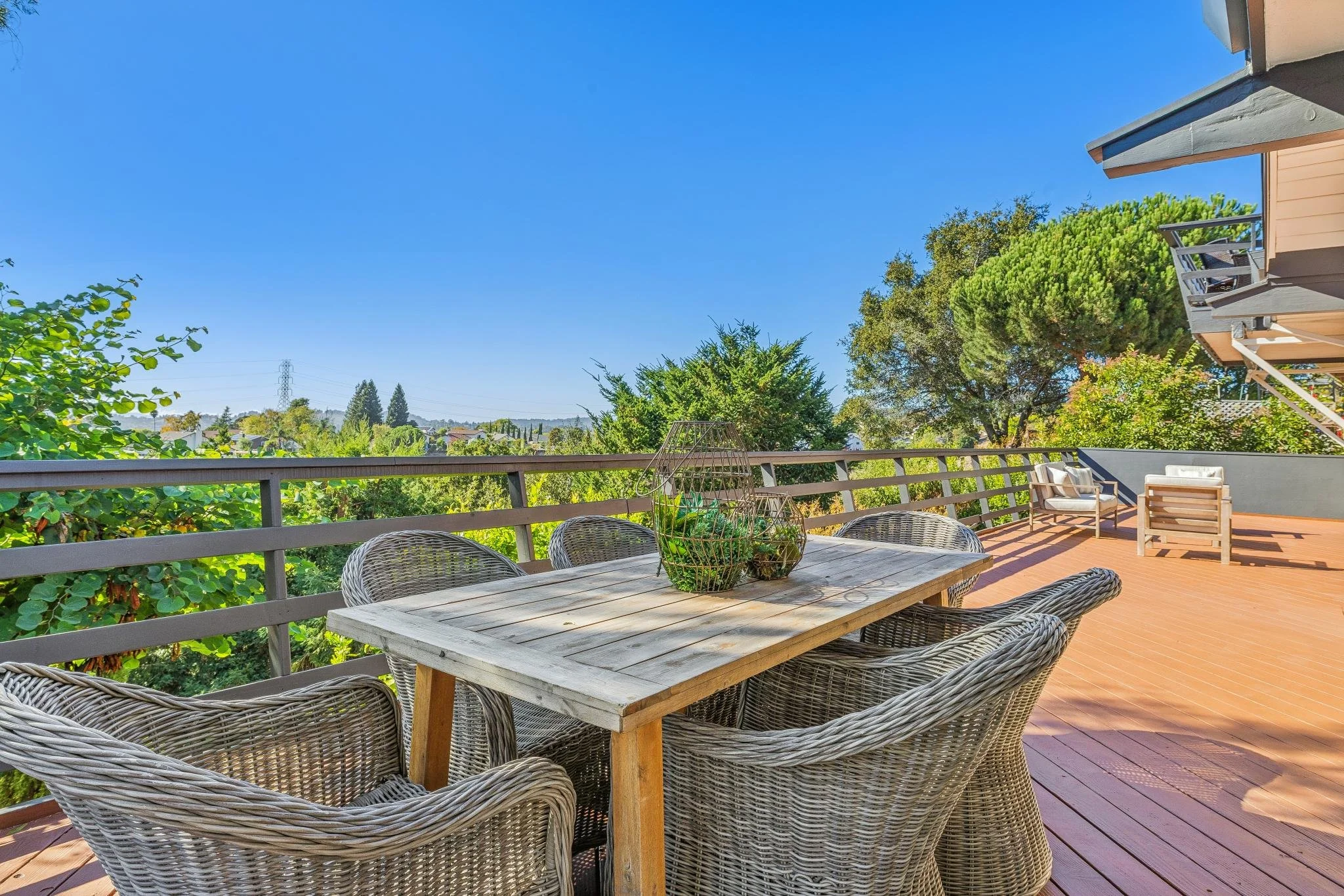 Outdoor wooden deck with a dining table and chairs, overlooking green trees and a clear blue sky.