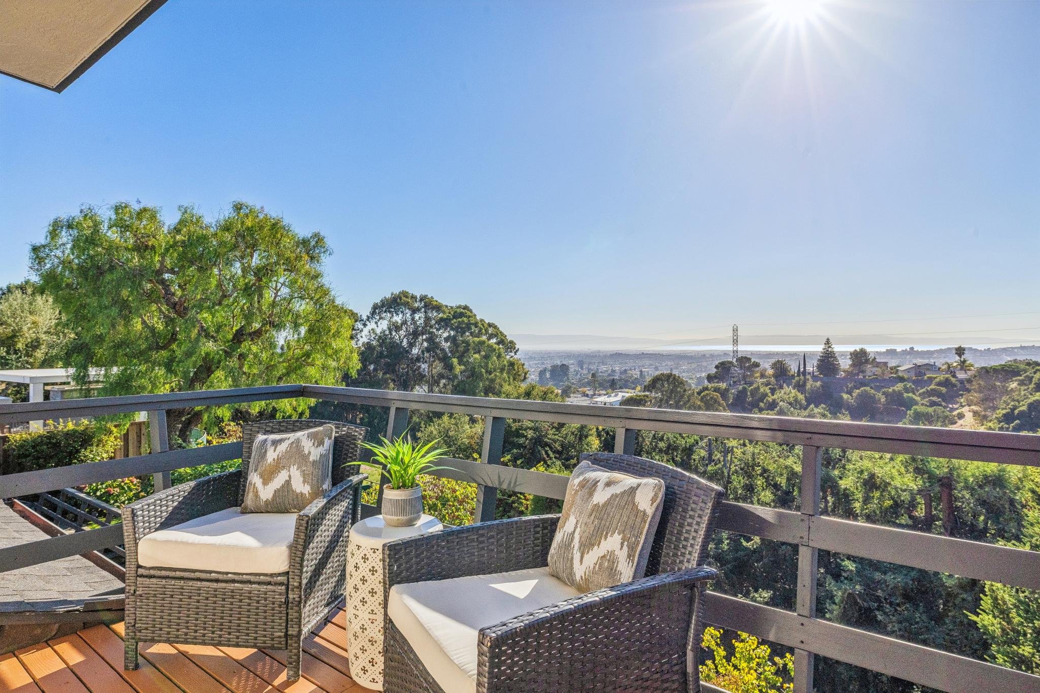 A balcony with wicker chairs and a small table holding a potted plant, overlooking trees, rooftops, and a body of water under a clear blue sky with the sun shining.