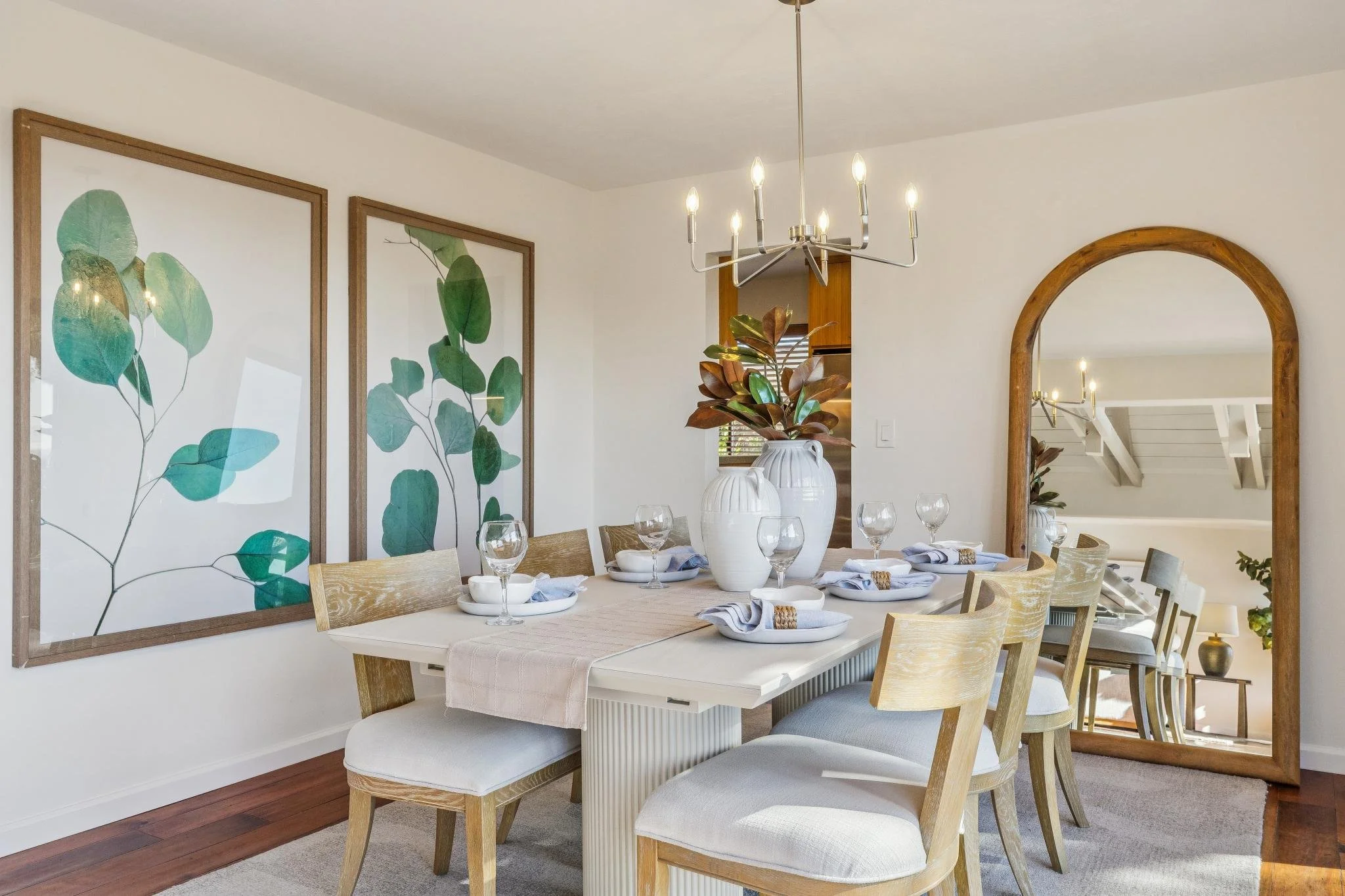 A dining room with a wooden table set with white and blue place settings, clear glasses, and white napkins. Two large white vases with brown and green leaves serve as a centerpiece. The room has framed botanical art and a wooden mirror, with natural 