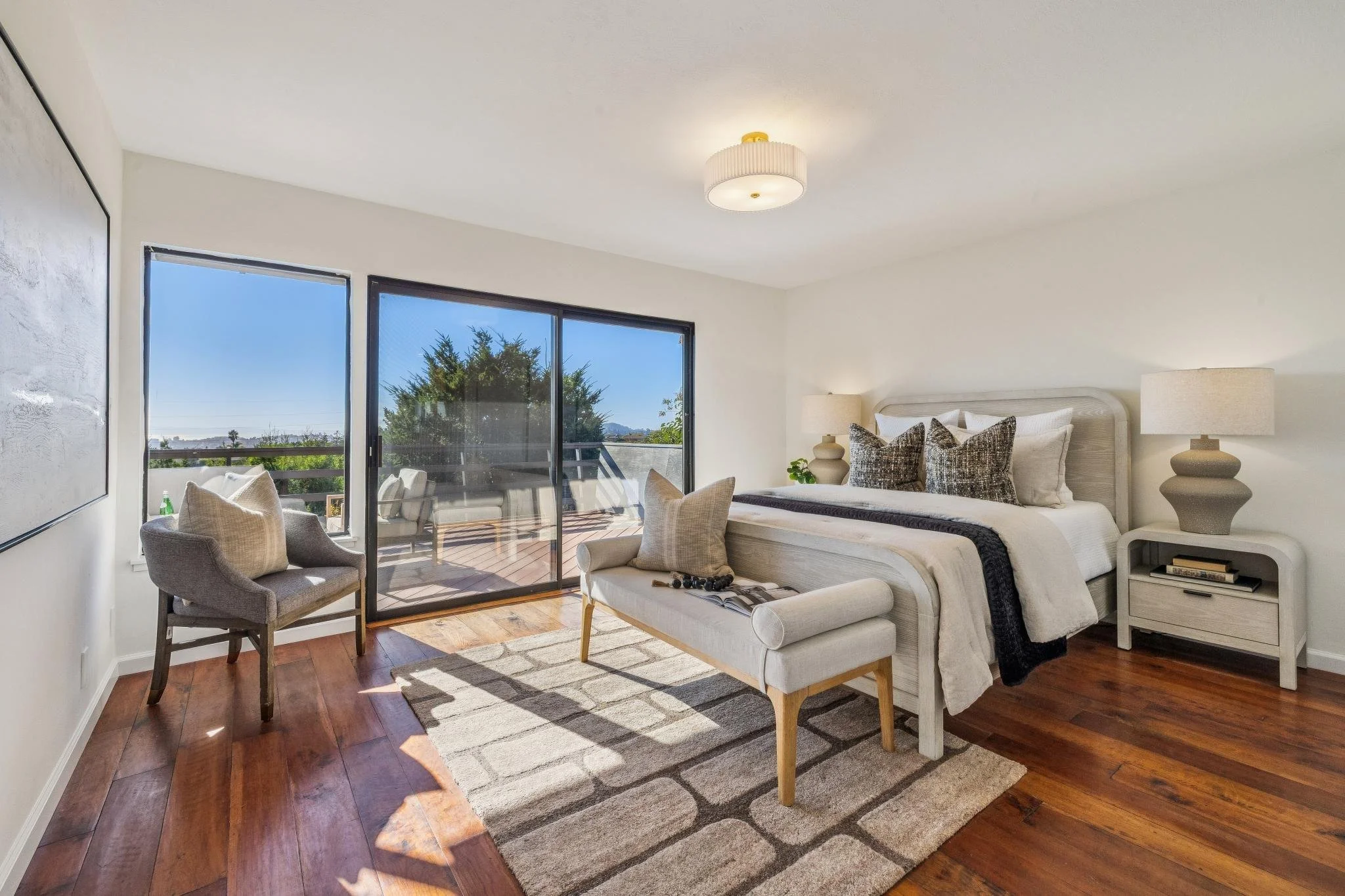 Bright bedroom with white walls, wooden floor, and large sliding glass door leading to a balcony with outdoor seating. The room has a white bed with pillows, bedside tables with lamps, and chairs near the window.