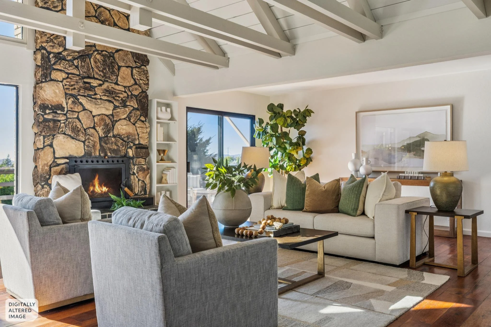 Living room with stone fireplace, white sofa, armchairs, green plants, side tables with lamps, large window showing outdoor trees, framed picture on wall, wooden floor, and area rug.