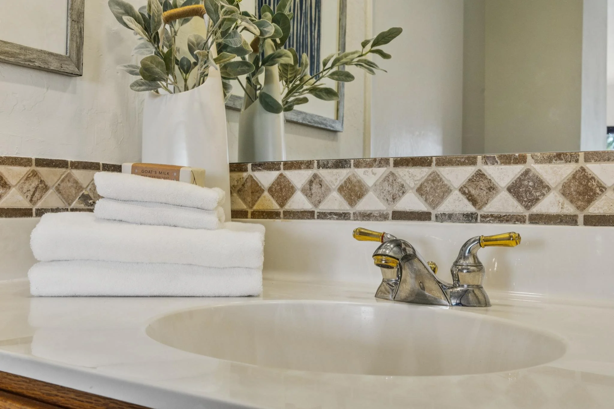A bathroom sink with a stack of three white towels, a soap bar labeled 'Goat's Milk', and a pair of yellow-handled faucets. Behind the sink, there are two white vases with green leafy plants, a mirror, and a tiled backsplash with diamond-shaped brown