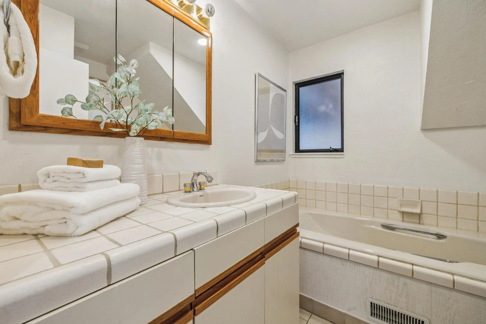A bathroom vanity with stacked white towels, a white vase with green foliage, a large mirror, and a window.