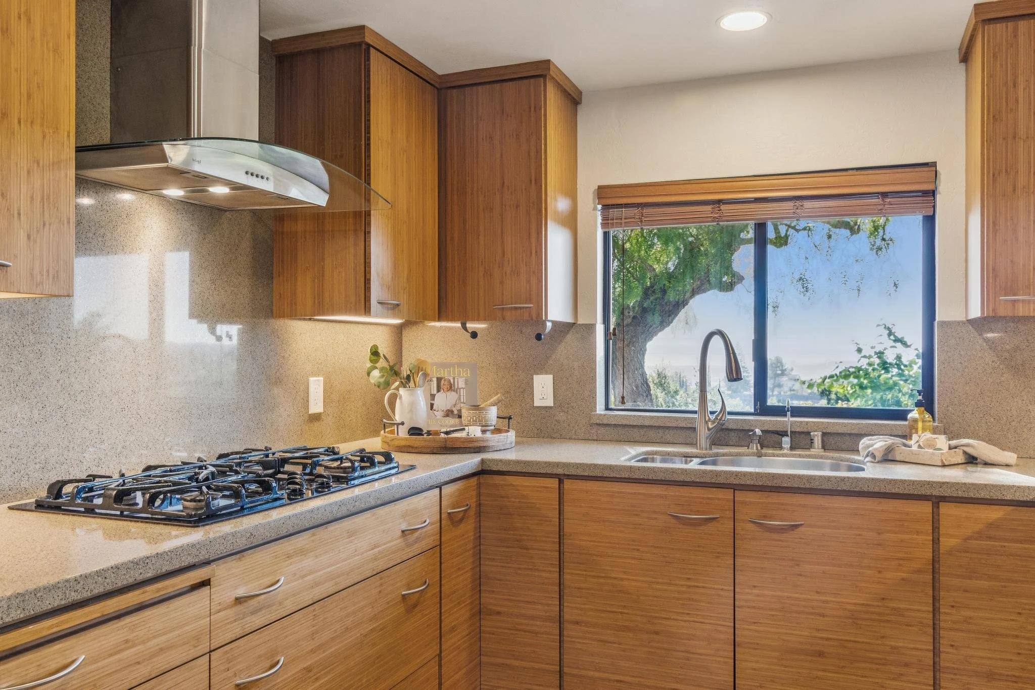 Kitchen with wood cabinets, a beige speckled countertop, a gas stove, a window with blinds showing a large tree outside, and some kitchen items like a vase, a book, and a dish towel.