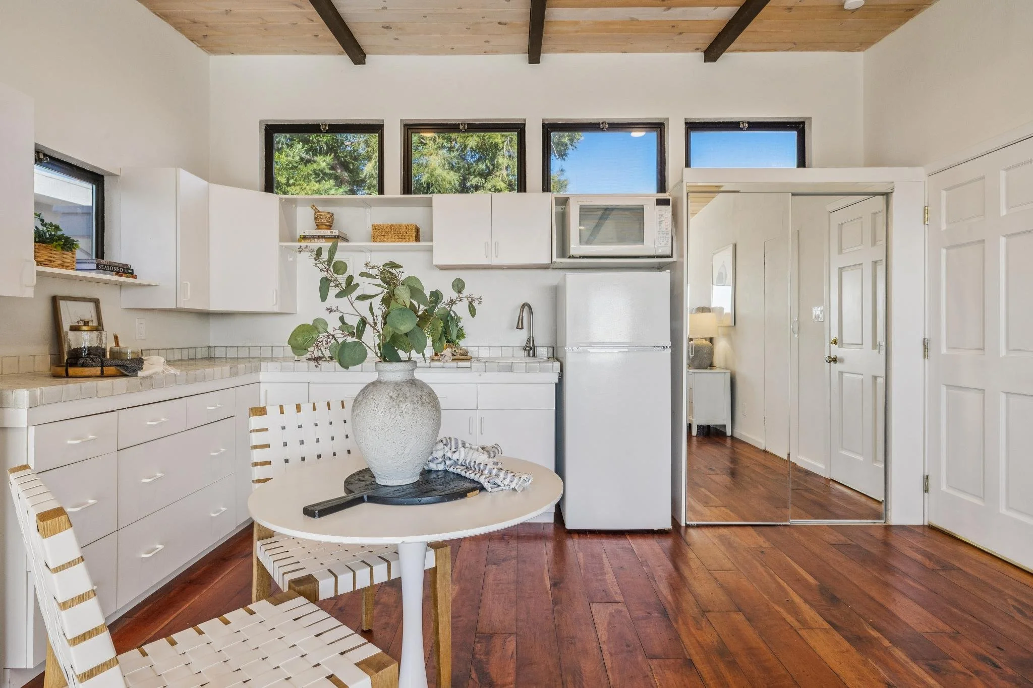 Kitchen with wood ceiling beams, white cabinets, a small round table with a vase, and a window with a view of trees.