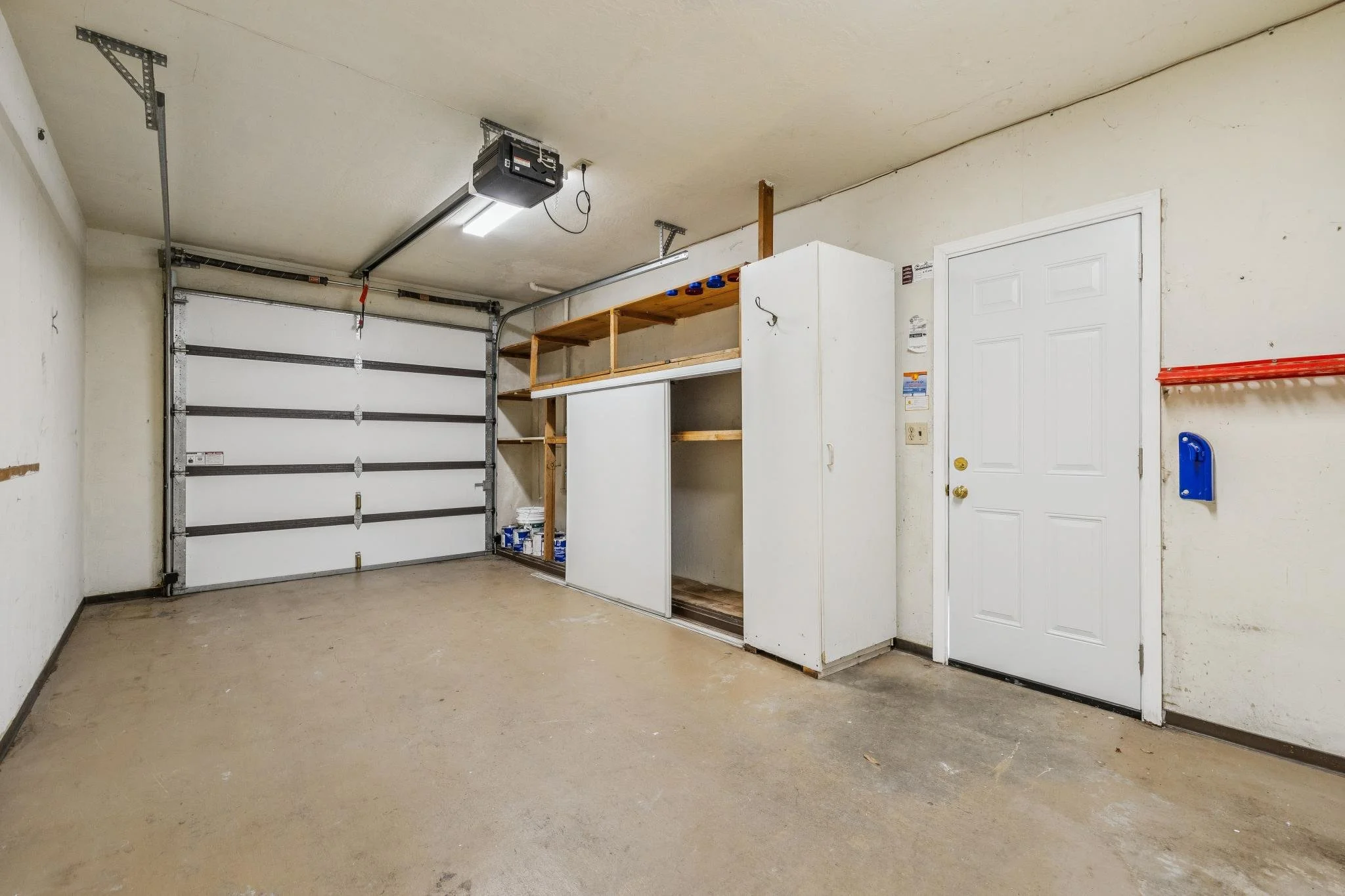 Empty garage with a closed garage door, white storage cabinets, and some shelving. The walls are plain with minor marks, and the floor is concrete with some dirt. Fluorescent lights illuminate the space.