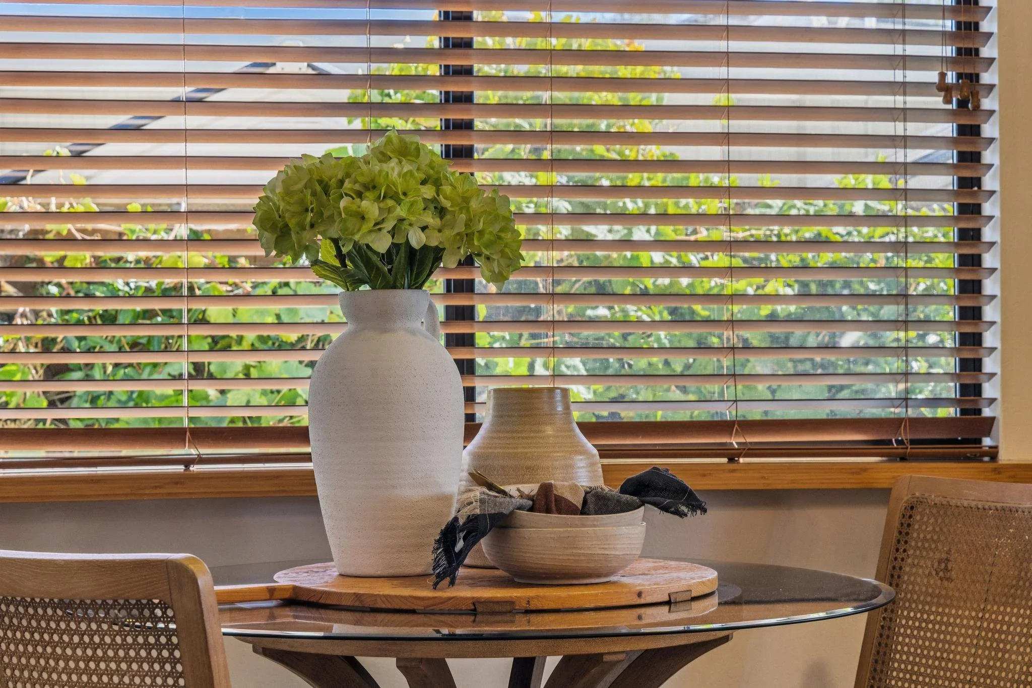A white vase with green hydrangea flowers and a beige decorative bowl on a wooden table in front of window blinds, with greenery outside.
