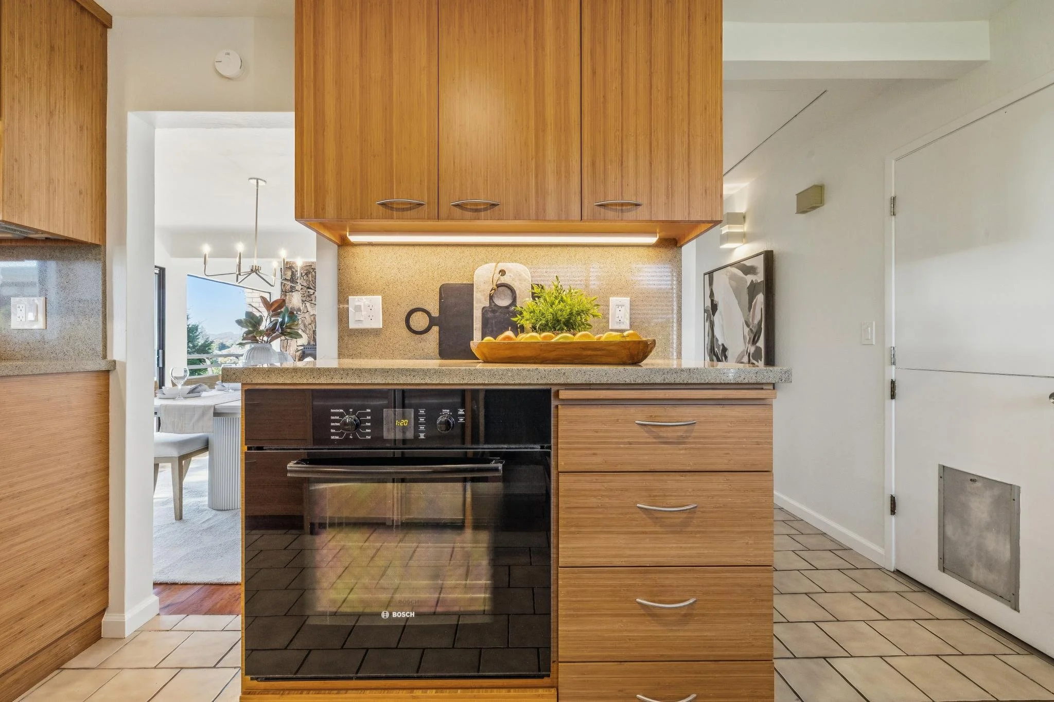  view of a kitchen with wooden cabinets, black oven, beige countertops, and decorative items including cutting boards, a potted plant, and fruit bowl.