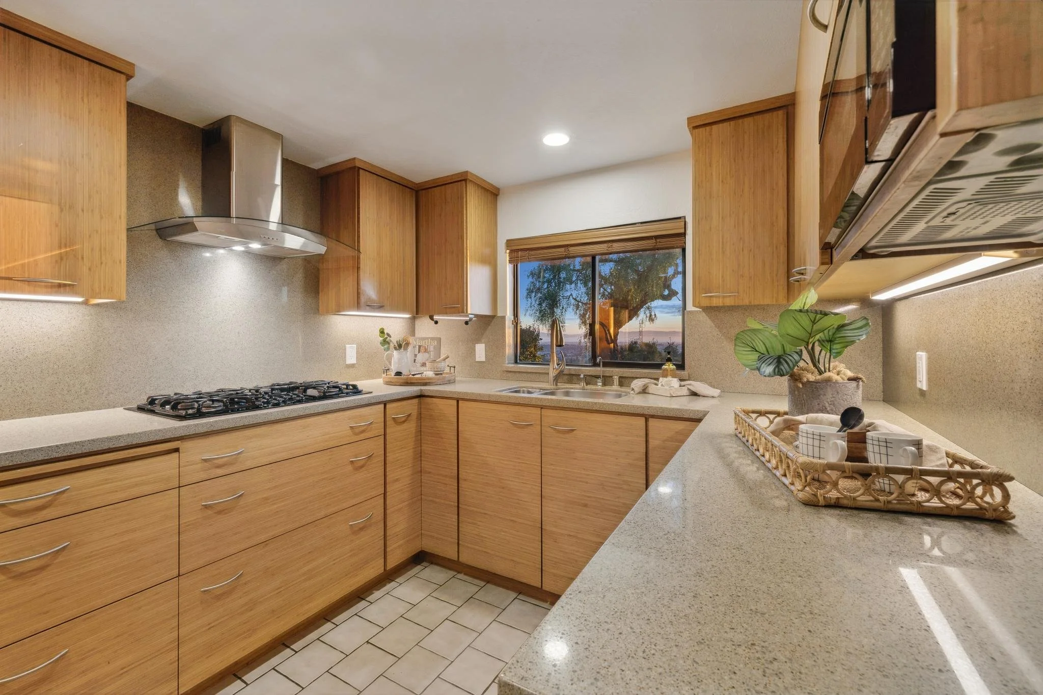 A modern kitchen featuring wooden cabinets, a beige speckled countertop, a stainless steel range hood, a gas stove, a window with a view of trees, a potted plant, and various kitchen items.