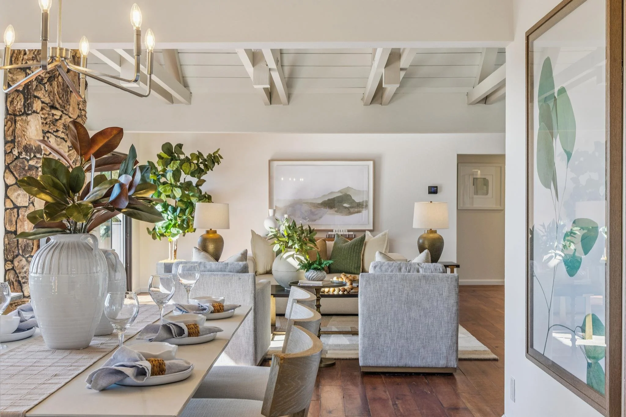 A cozy living room with a dining area, featuring a wood-burning stone fireplace, indoor plants, a chandelier, and a large wall art print of green leaves.
