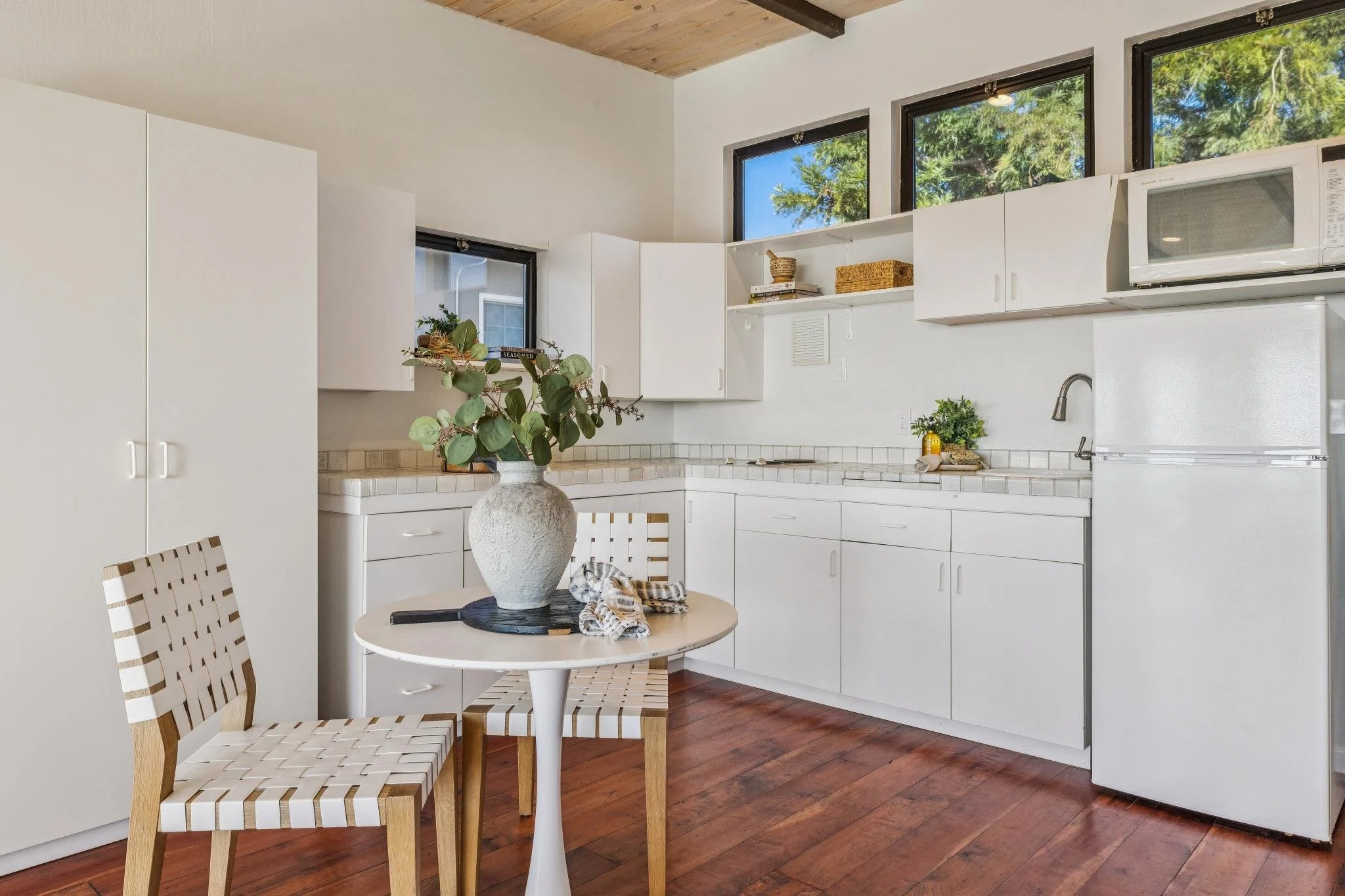 A white kitchen with cabinets, a microwave, and a small round table with woven chairs. There is a large vase with greenery on the table. The kitchen has upper windows letting in natural light.