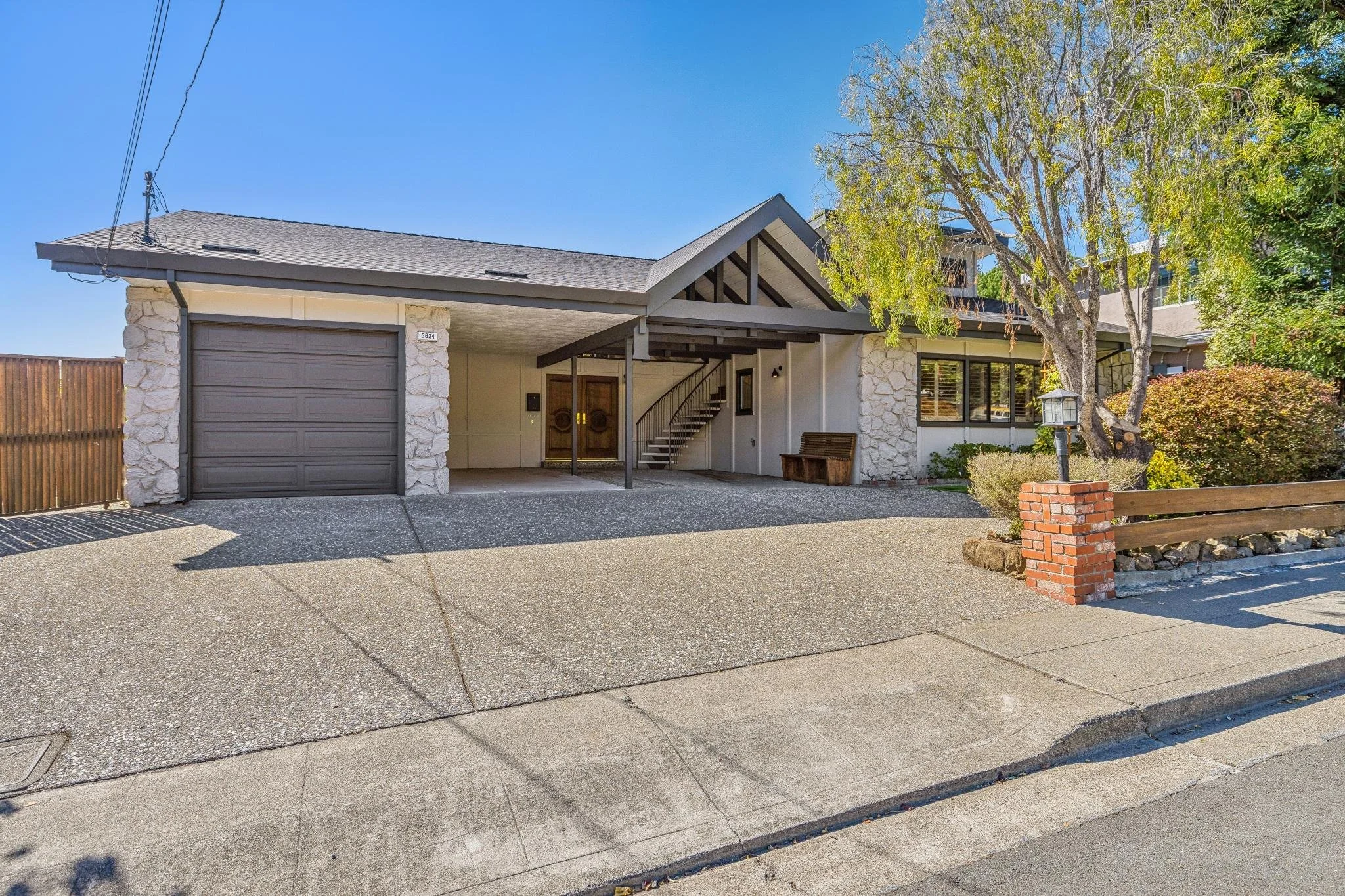 Front view of a modern house with a garage, stone and wood exterior, and a large tree in the yard under a clear blue sky.