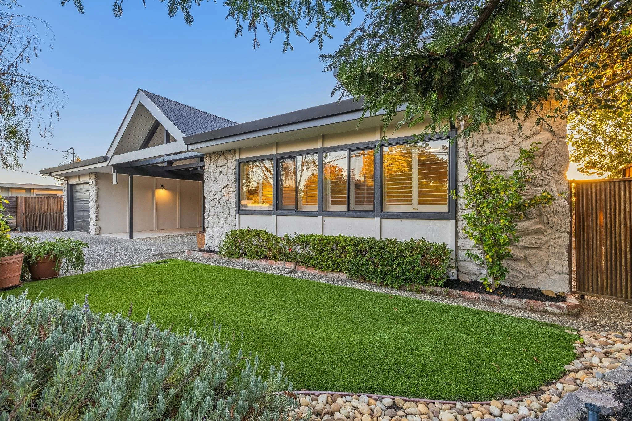 A modern house with large windows, stone facade, and a gabled roof, surrounded by a well-maintained lawn, shrubbery, and trees at sunset.