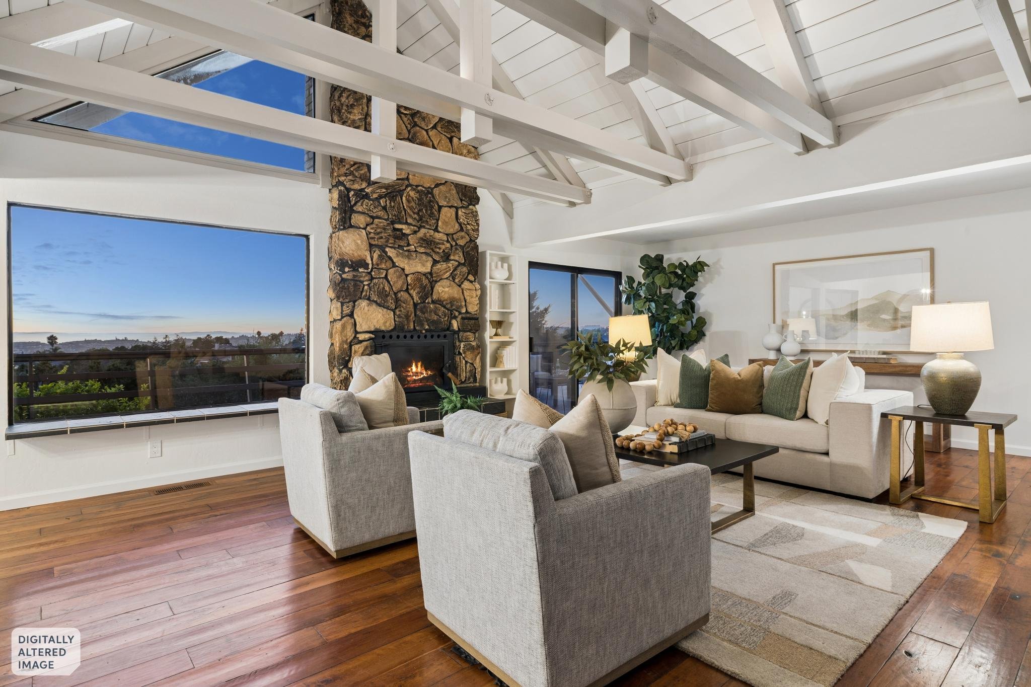 Living room with large windows, white ceiling beams, a stone fireplace, beige sofa, armchair, and various decorative items. View of a clear blue sky outside.