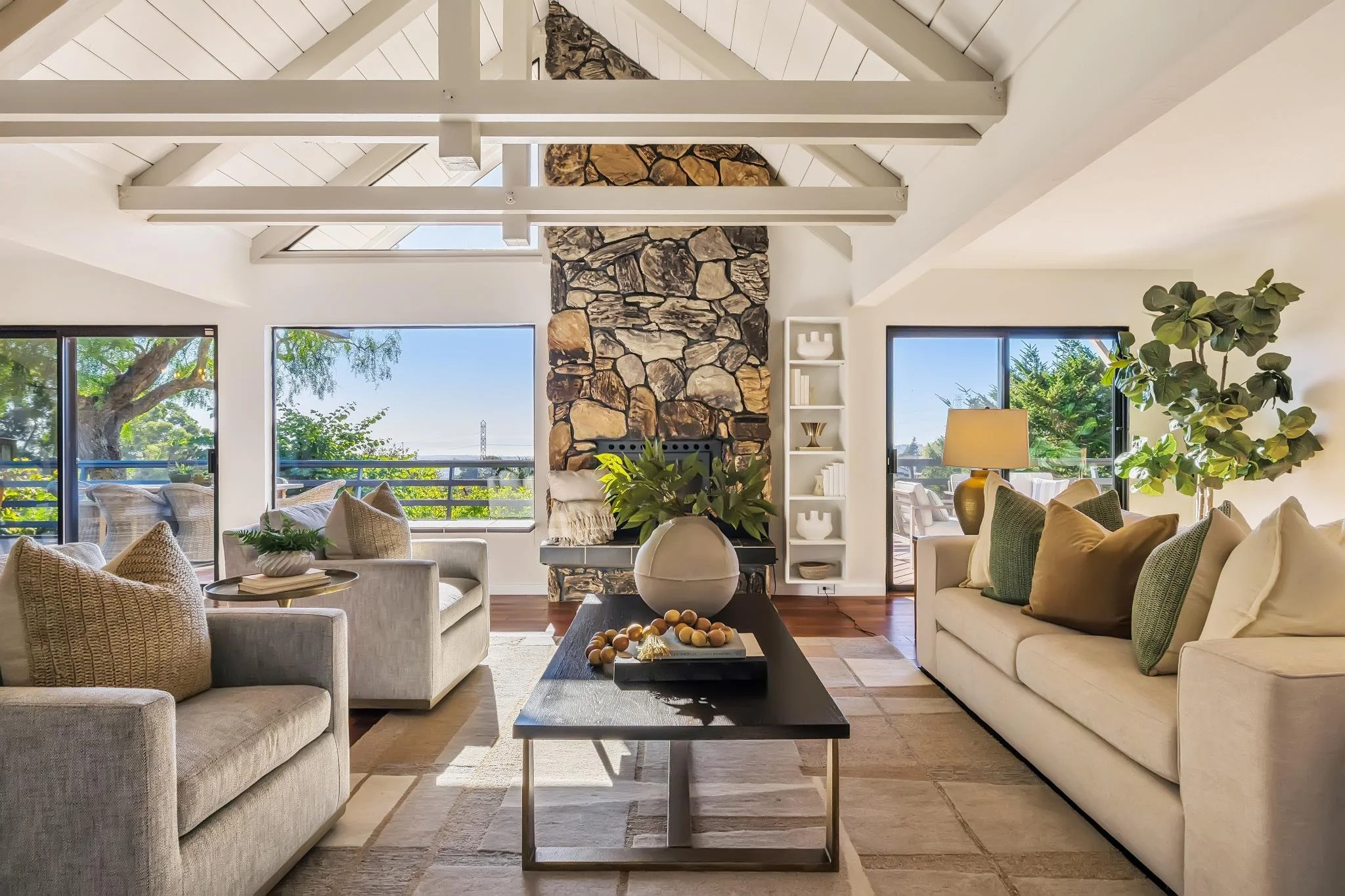 Living room with large windows, a stone fireplace, and high vaulted ceiling with white beams. Furniture includes beige sofas with green and beige pillows, wicker armchairs, a dark coffee table with decorative items, and a white bookshelf. There is a 