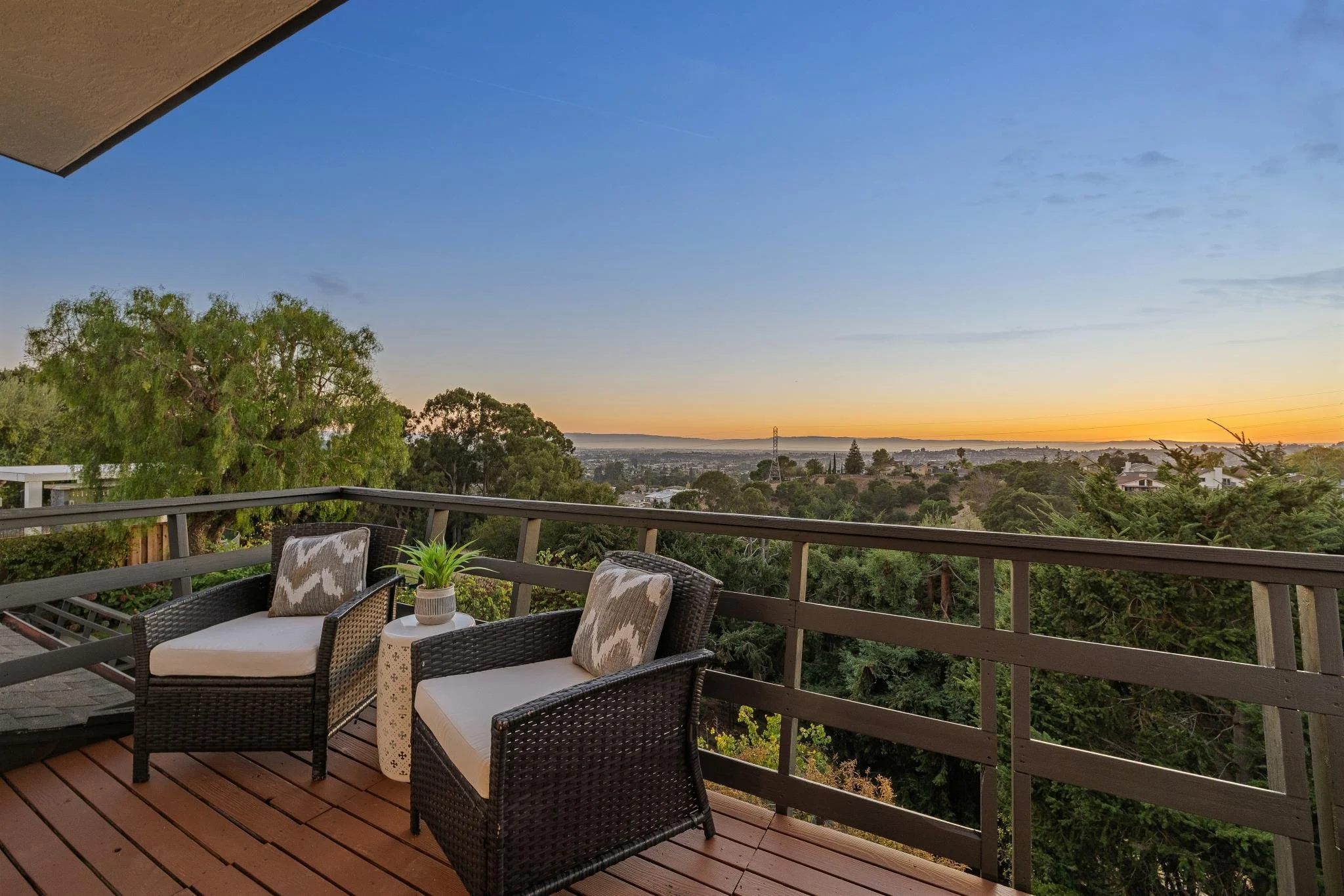 A balcony with patio furniture, including two cushioned chairs, a small table, and a potted plant, overlooking a scenic view of trees, houses, and a sunset sky.