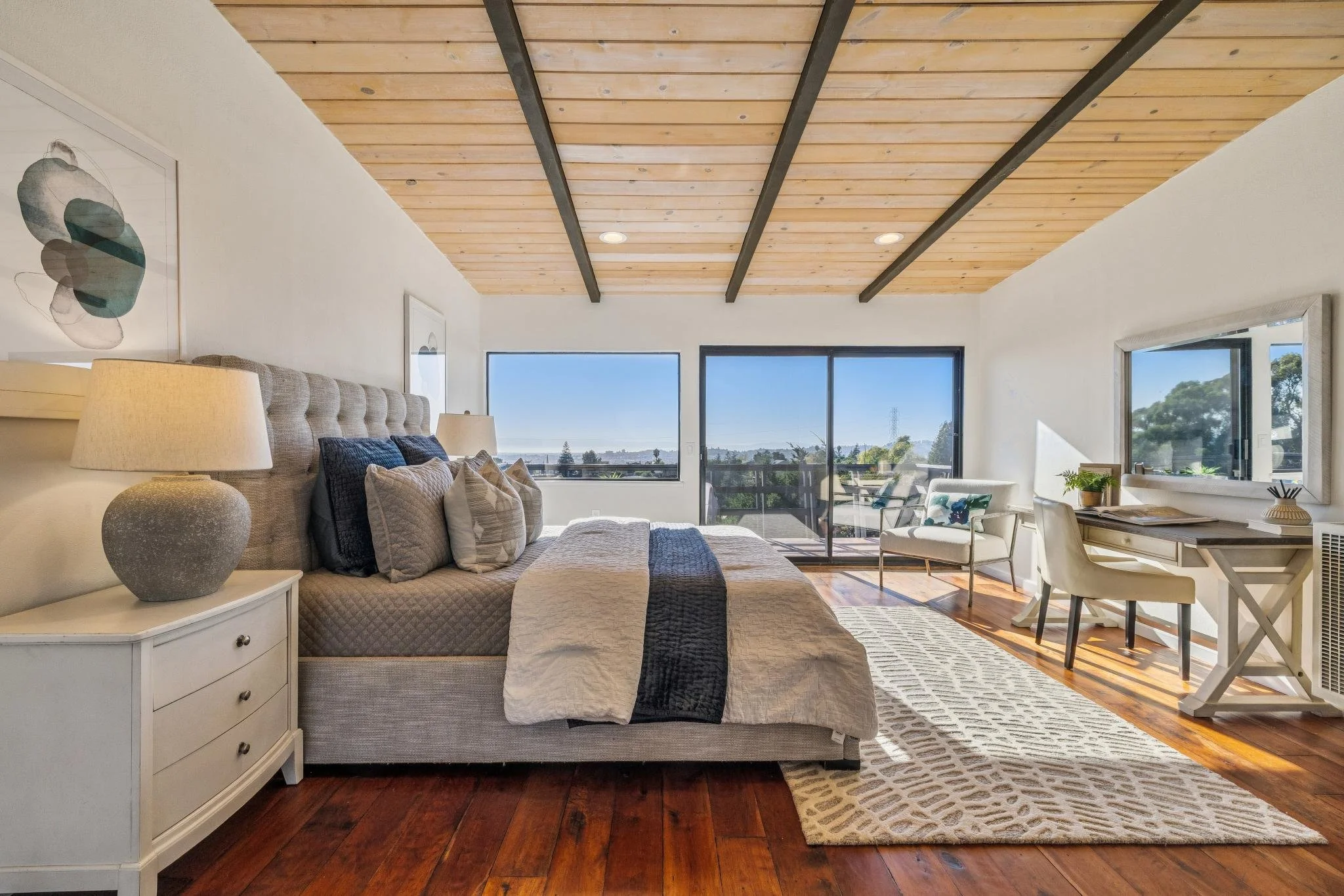 Living room with a beige tufted sofa, a white side table with a textured lamp, large windows showing a view outside, a wooden ceiling with black beams, a patterned rug on hardwood flooring, and chairs near a desk with a small plant.