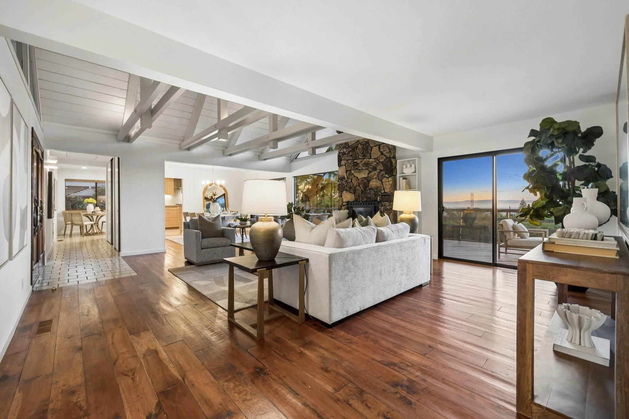 Living room with white ceiling and exposed beams, hardwood floors, white and gray furniture, large window with view of trees and a sunset, and decorative vases.
