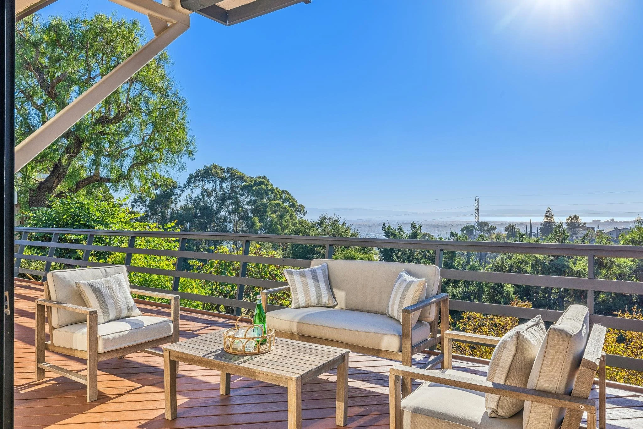 Outdoor patio area with wooden furniture including sofas and chairs with cushions, a small table with a tray holding a green glass bottle, overlooking trees, hills, and a clear blue sky.