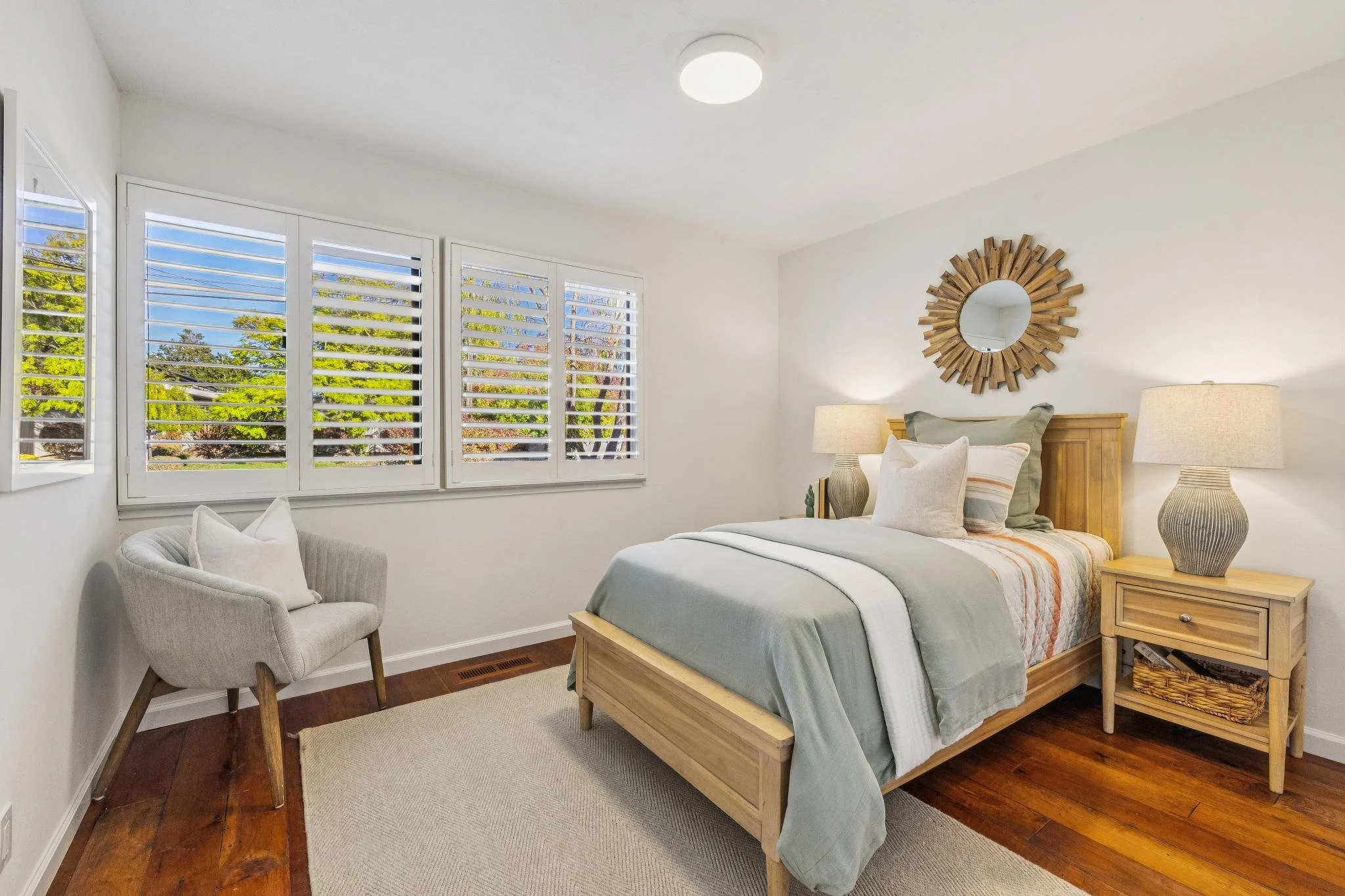 A bedroom with white walls and wooden floors, featuring a large bed with light-colored bedding and pillows, a wooden nightstand with a vase and a lamp, a white armchair, and a mirrored sunburst wall decoration, with large windows with white shutters 