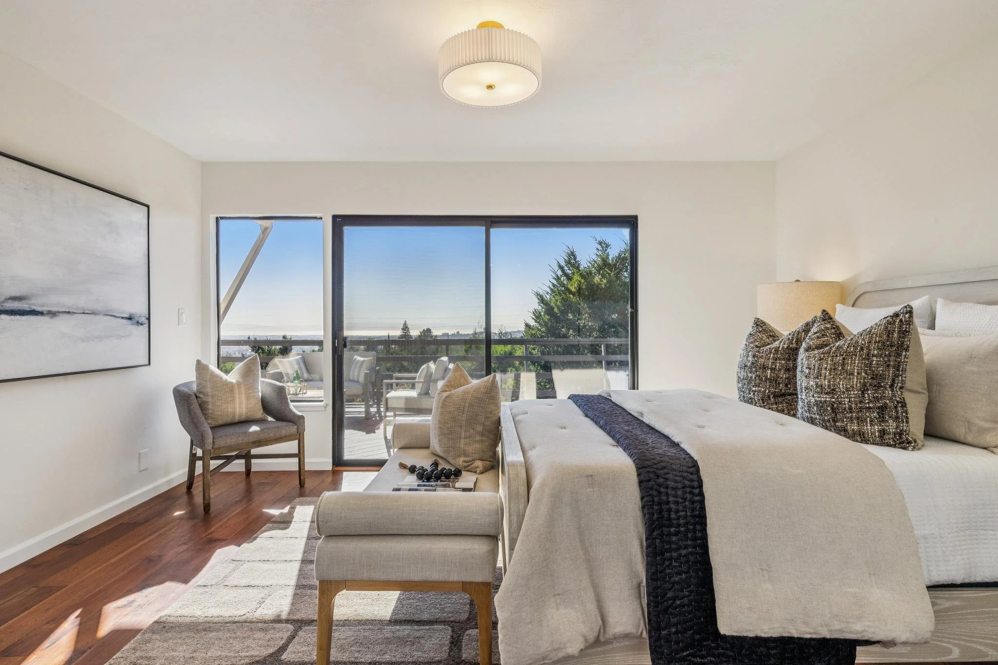 Living room with beige sofa, pillows, and a black blanket, large glass sliding door leading to balcony with outdoor seating, abstract painting on wall, lamp, and wooden floor with rug.