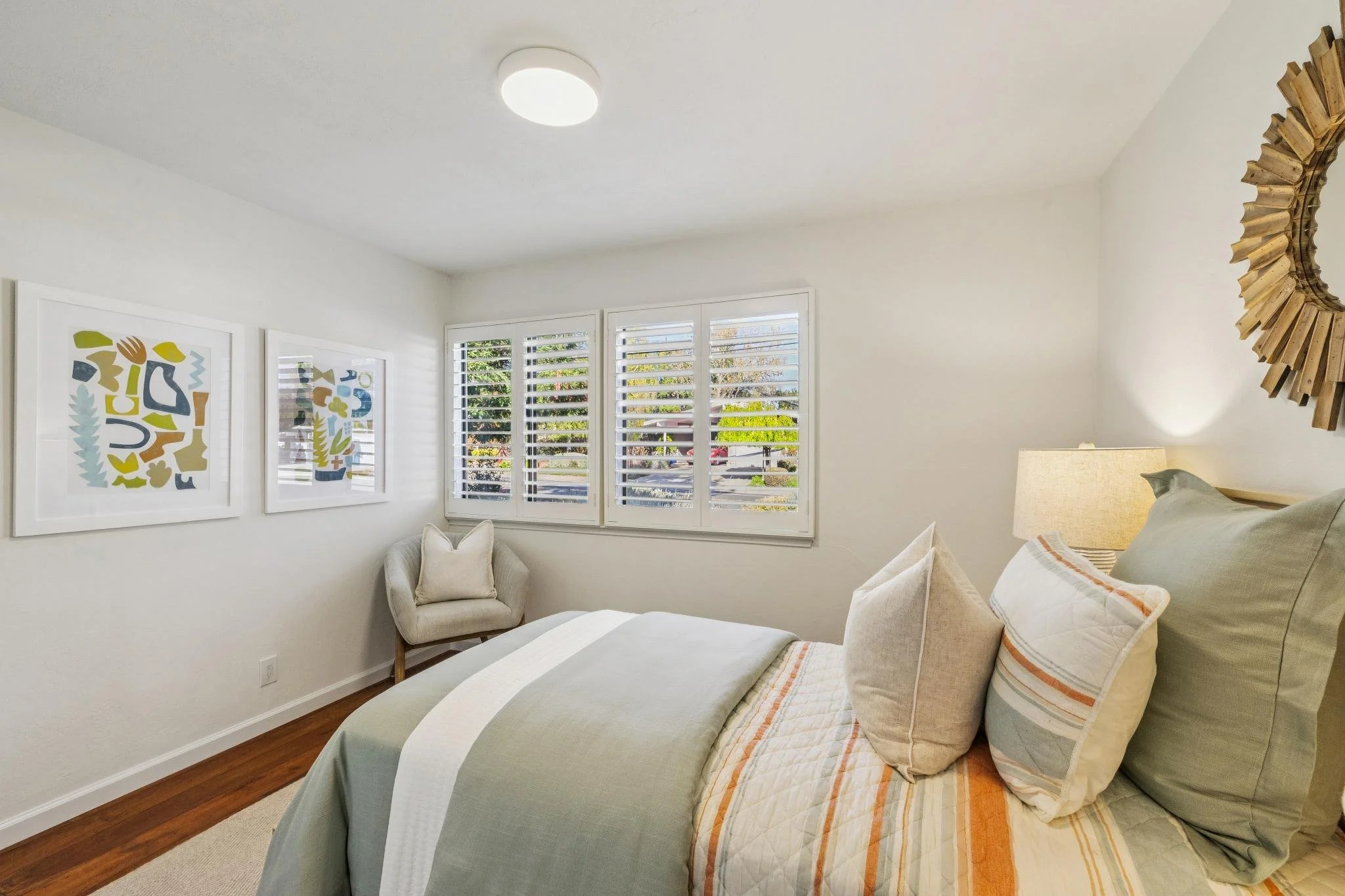 A cozy bedroom with a cream-colored armchair near a window with white shutters. The bed has a light green quilt with orange stripes and several pillows. There is a bedside lamp, and the wall above the bed has a wooden sunburst mirror. Two framed abst