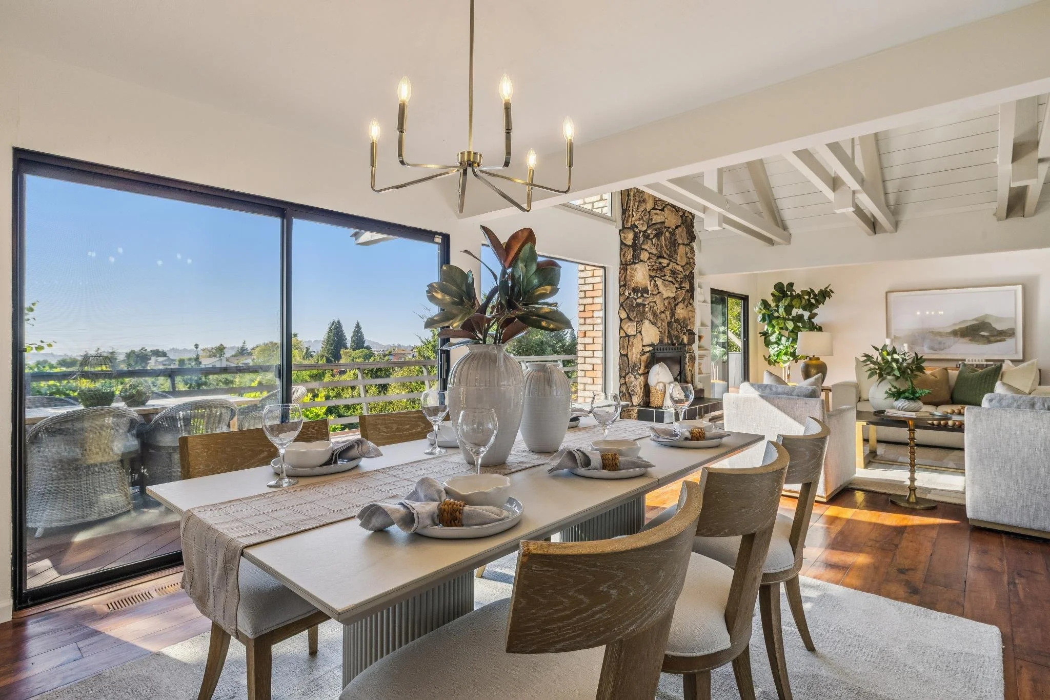 Modern living room with large windows, stone fireplace, ceiling chandelier, and dining table set for a meal with vases, glasses, and plates.