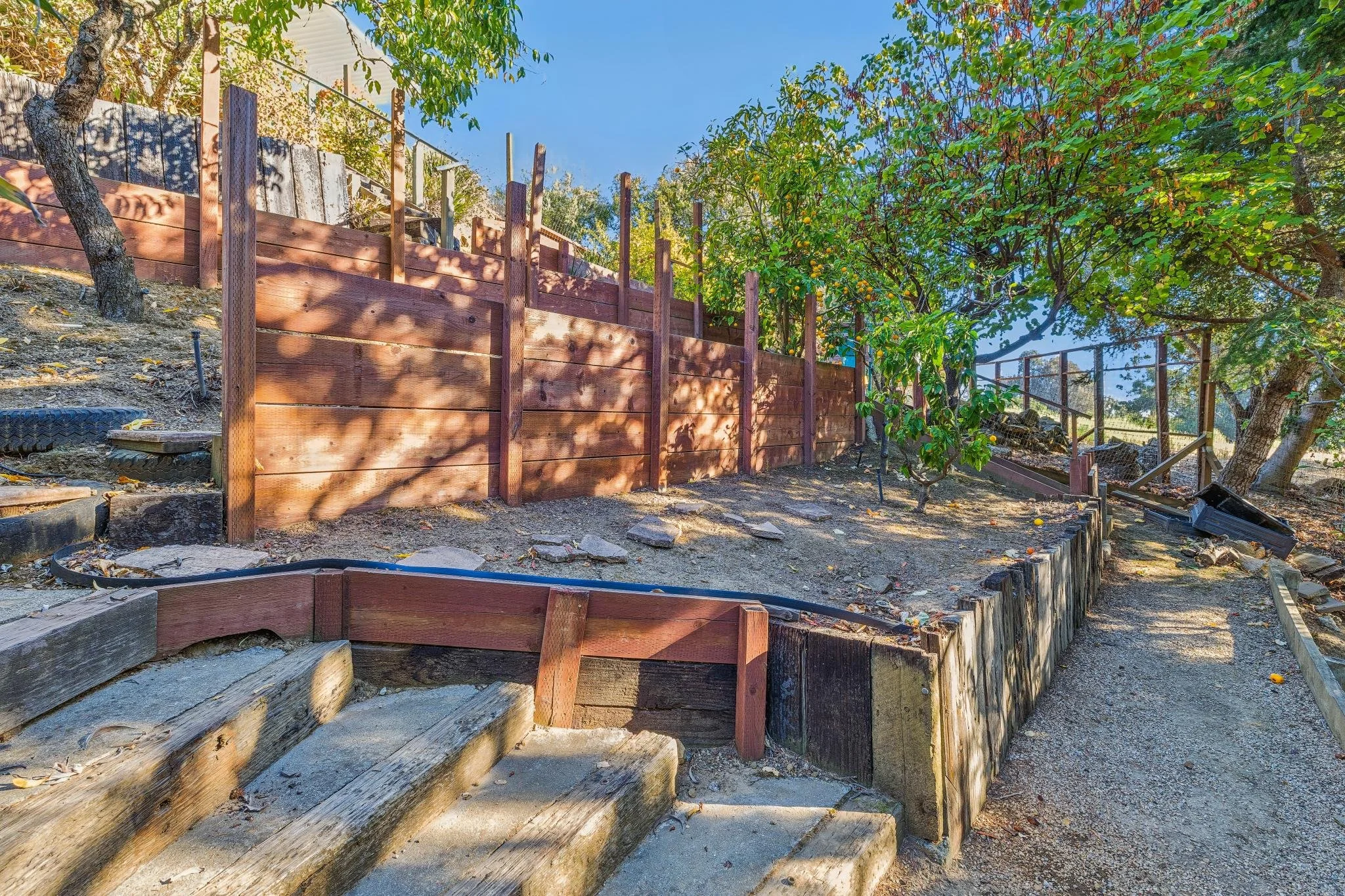 A backyard garden area with wooden retaining wall, stone steps, green trees, and a clear blue sky.