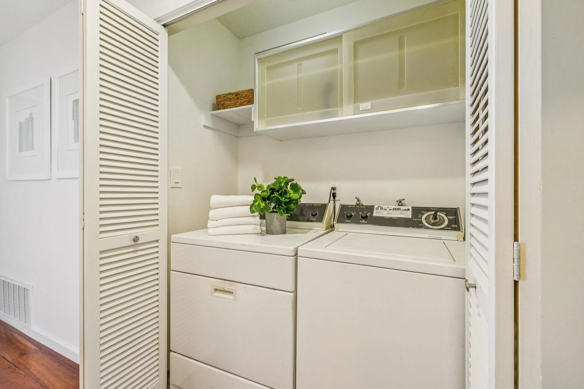 A laundry area with a washing machine and dryer, a plant in a gray pot, neatly folded towels, and storage cabinets, within a closet space with white louvered doors.