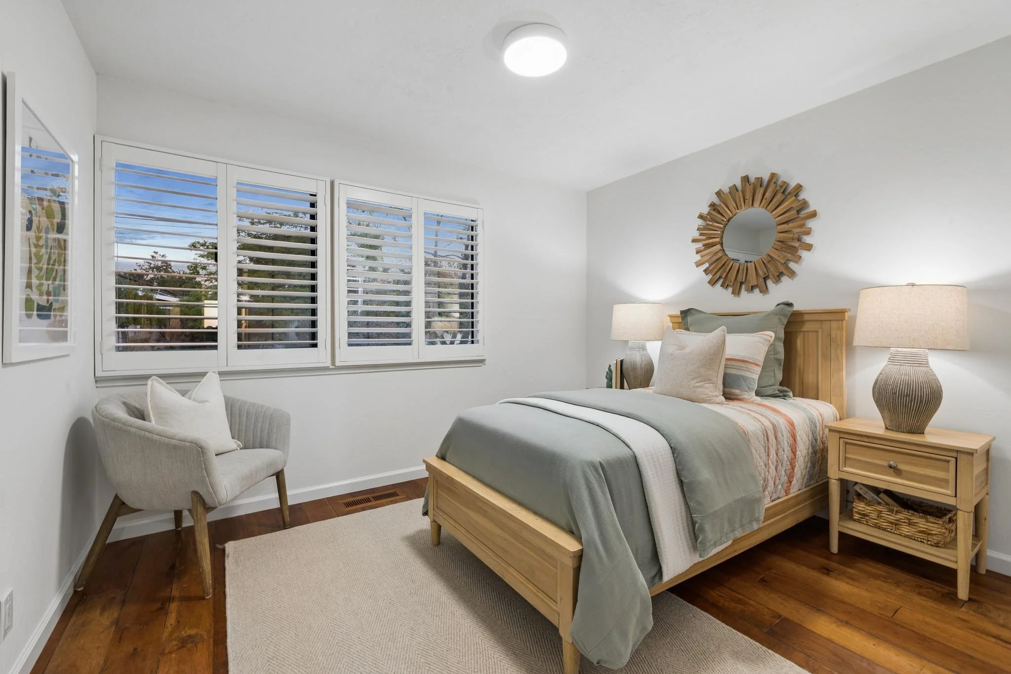 A cozy bedroom with a wooden bed frame, beige bedding, and a striped quilt. There are matching beige lamps on wooden nightstands, a decorative round mirror made of wooden slats above the bed, a white armchair with pillows near large windows with whit