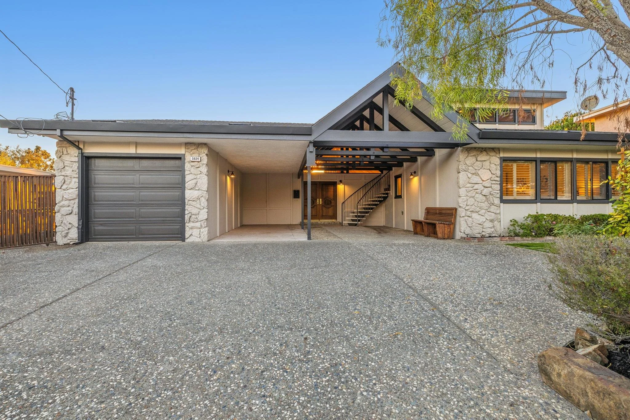 Front view of a modern house with a garage, stone and white exterior walls, a covered entrance with a staircase inside, and landscaping with bushes and a tree, during sunset.